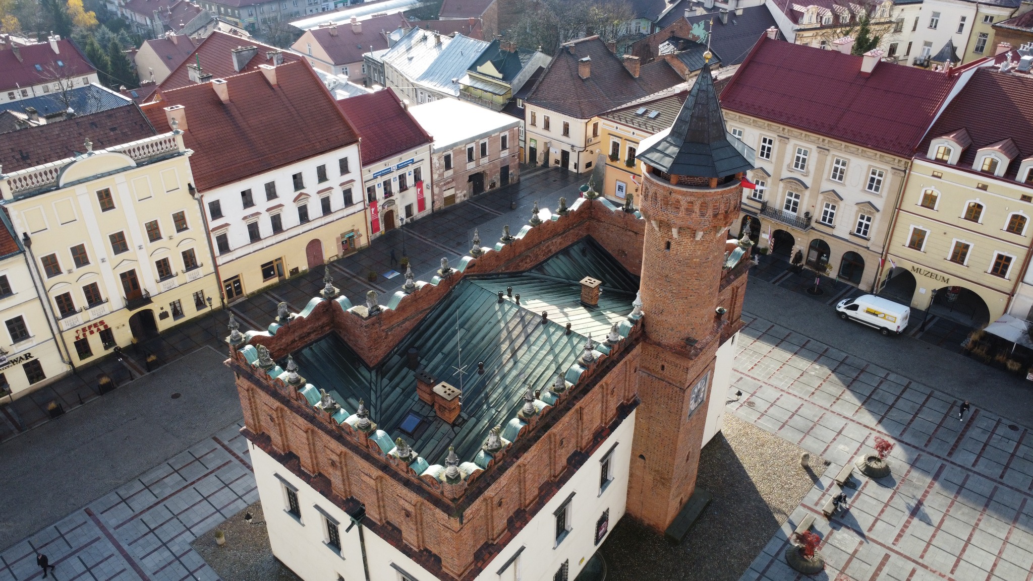 Widok z góry na renesansowy ratusz z cegły z wysoką wieżą, otoczony zabudową miejską i placem z geometrycznym wzorem kostki.