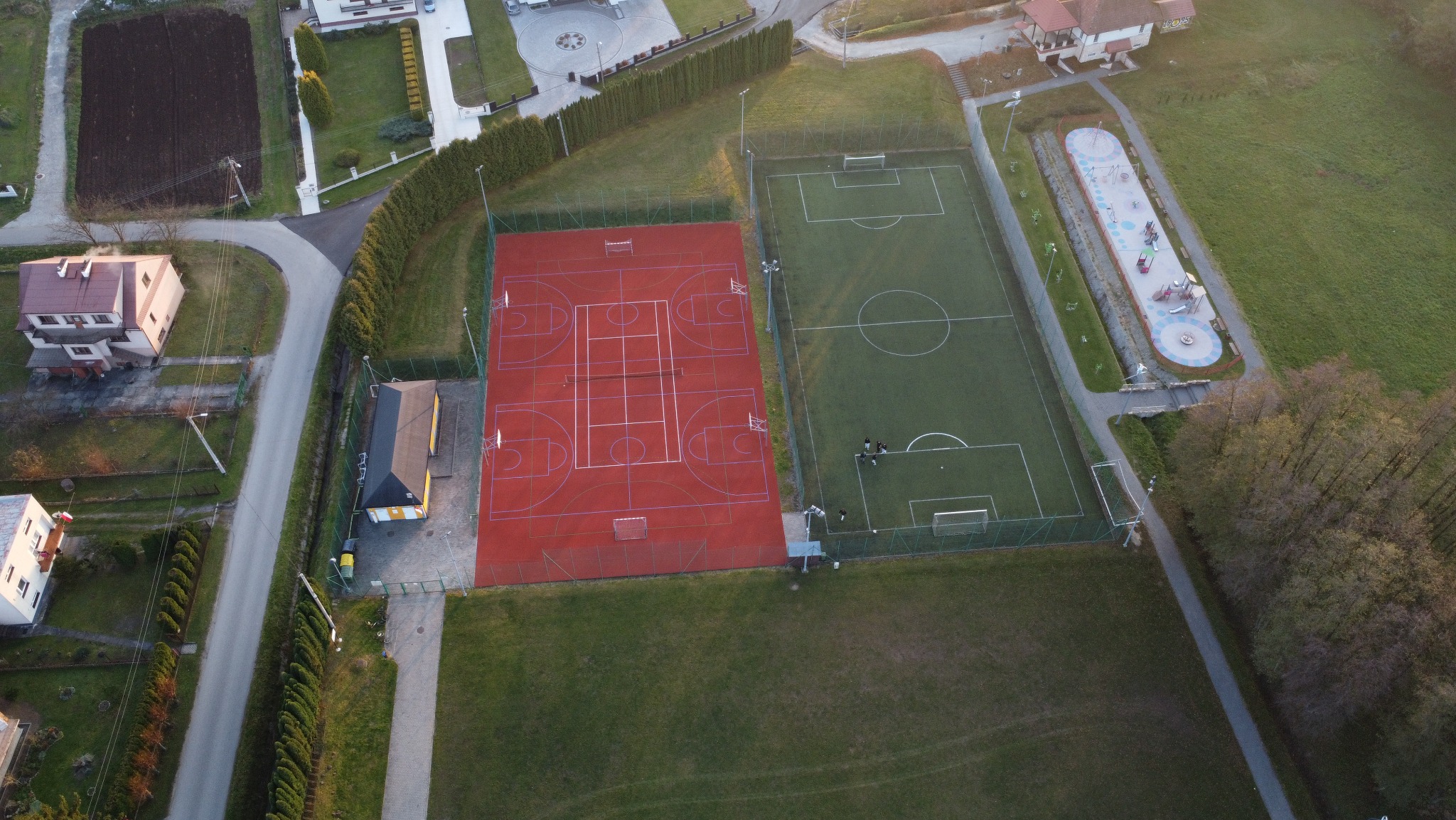 Widok z lotu ptaka na kompleks boisk sportowych: czerwone boisko do koszykówki i zielone boisko do piłki nożnej, obok plac zabaw dla dzieci, otoczone zielenią i zabudową mieszkalną.