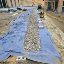 Wykop wyłożony niebieską tkaniną, wypełniony kamieniami drenażowymi wzdłuż budynku. W tle ziemia umocniona siatką i sprzęt budowlany.