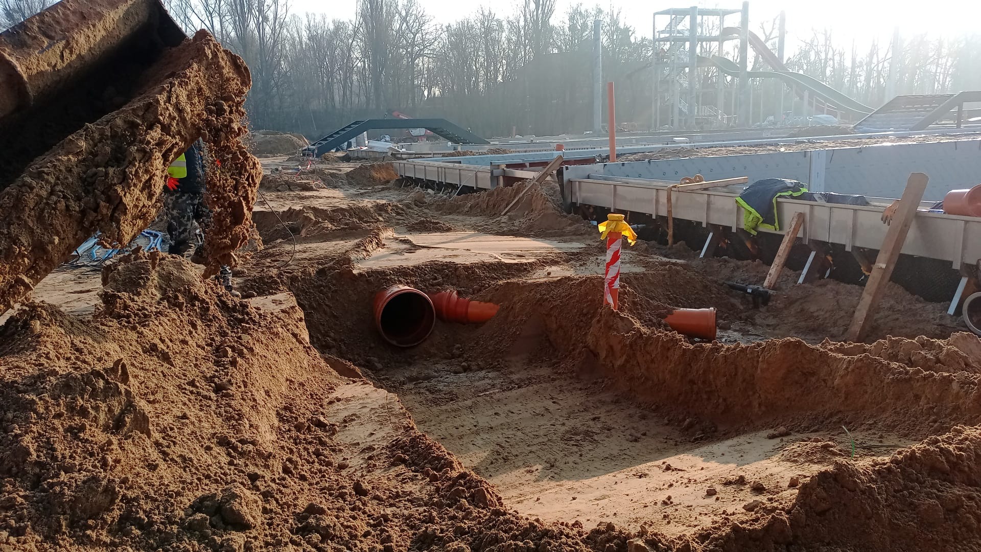 Wykopy pod rury kanalizacyjne na placu budowy, widoczne rury w wykopach, w tle konstrukcja budowlana i drzewa, ziemia piaszczysta, częściowo w cieniu, częściowo oświetlona słońcem.