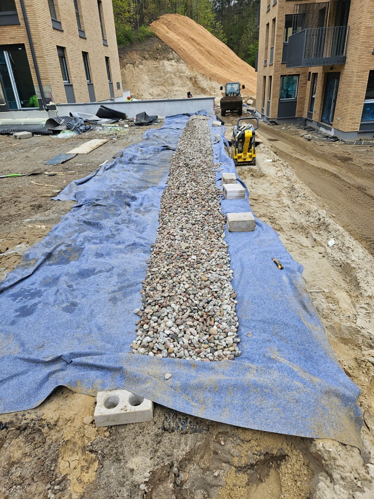 Wykop wyłożony niebieską tkaniną, wypełniony kamieniami drenażowymi wzdłuż budynku. W tle ziemia umocniona siatką i sprzęt budowlany.