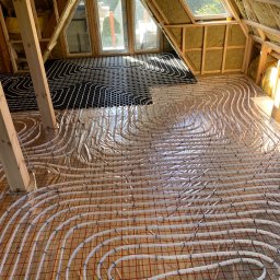 Rafał Jarząbek - Podłogówka w trakcie montażu na poddaszu: białe rurki układane spiralnie na folii z czerwoną siatką, część instalacji w czarnym kolorze. Widoczne okna dachowe i drewniana konstrukcja.