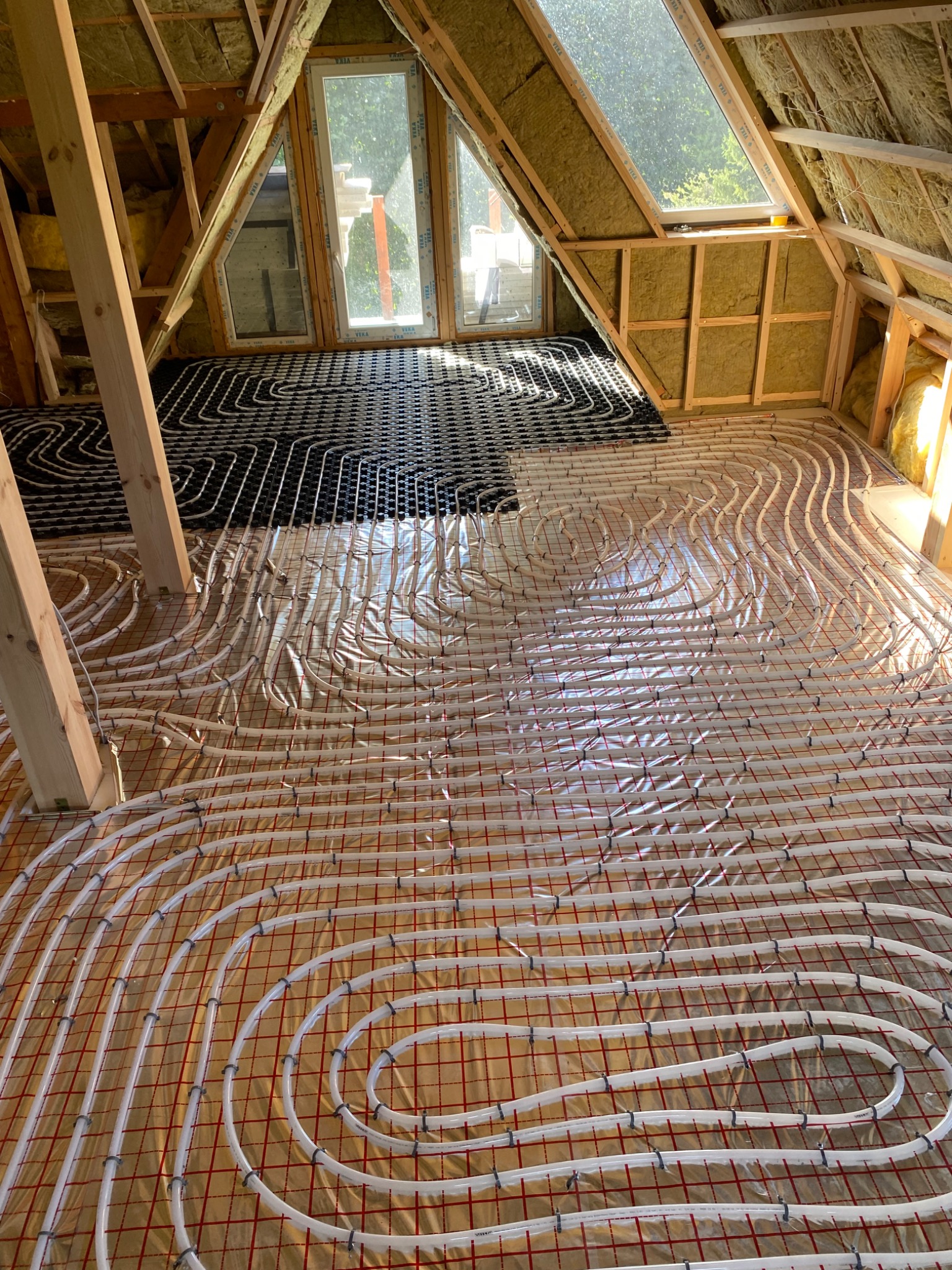 Podłogówka w trakcie montażu na poddaszu: białe rurki układane spiralnie na folii z czerwoną siatką, część instalacji w czarnym kolorze. Widoczne okna dachowe i drewniana konstrukcja.