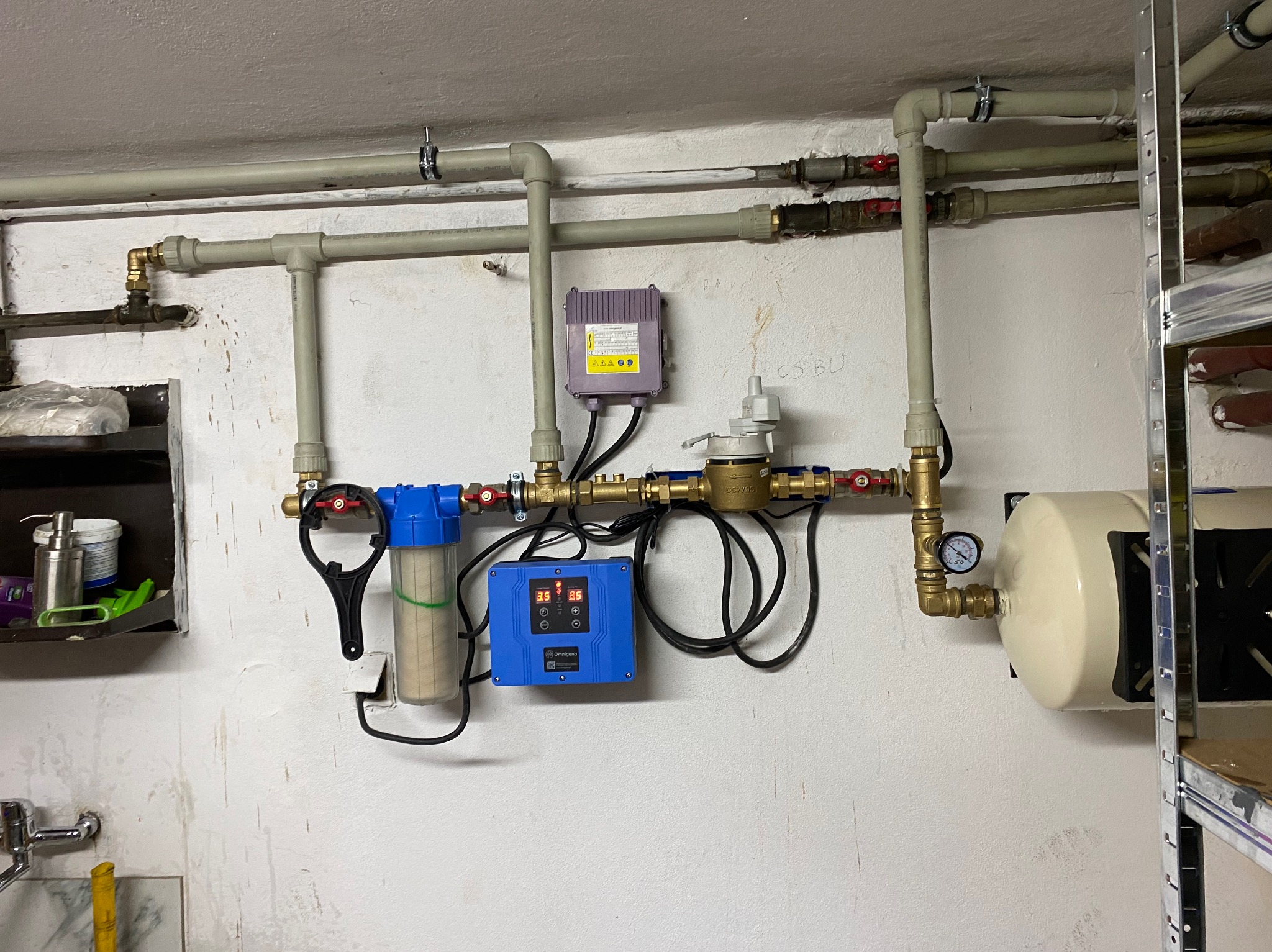Kompleksowa instalacja wodna z filtrem, sterownikiem i zbiornikiem wyrównawczym na ścianie. Widoczne rury, zawory i manometr. Realizacja instalacji hydraulicznej.