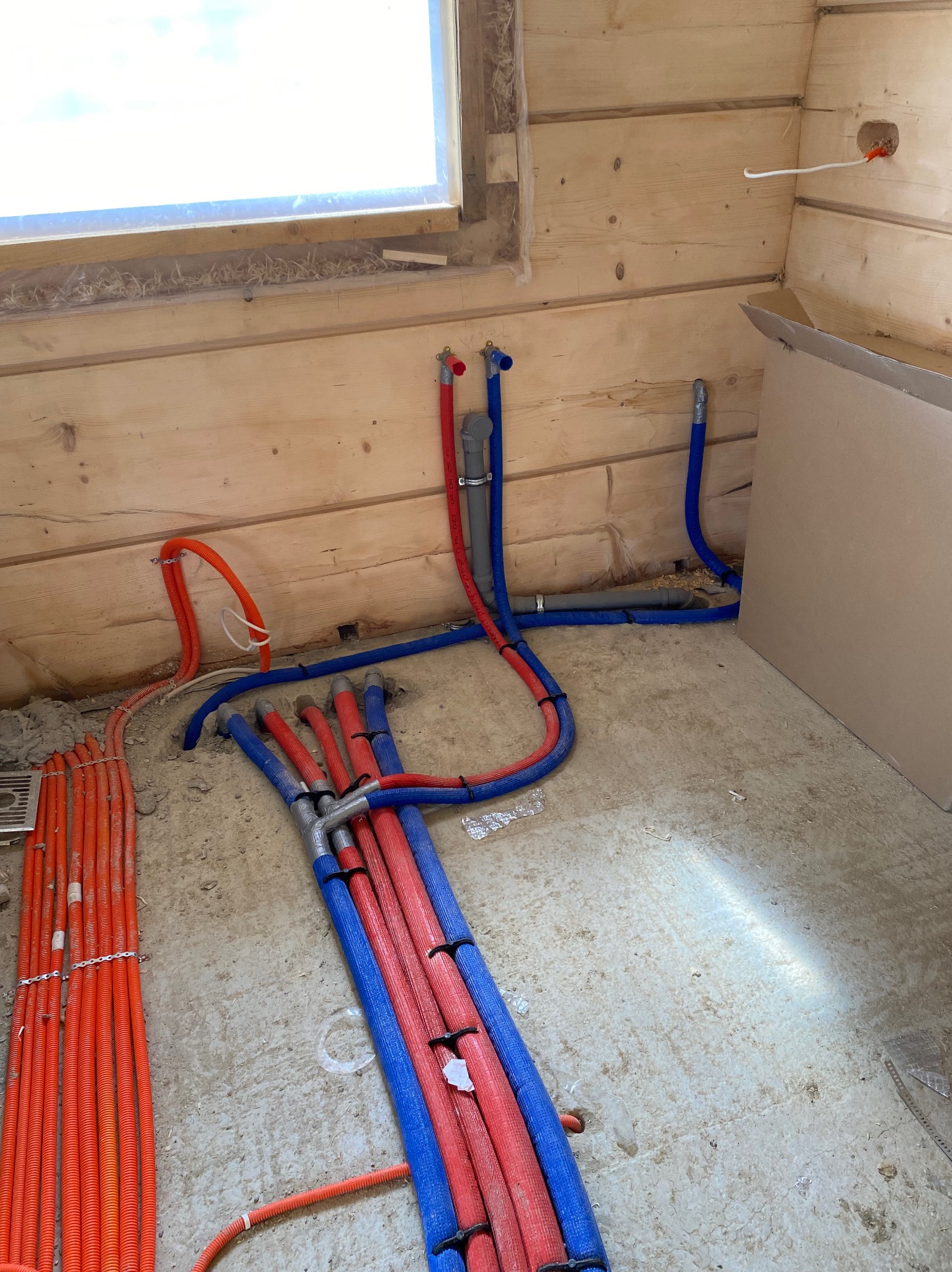 Instalacja ogrzewania podłogowego w drewnianym domu: rury w izolacji czerwonej i niebieskiej, częściowo zalane w szlichtę, widoczne okno i drewniane ściany.