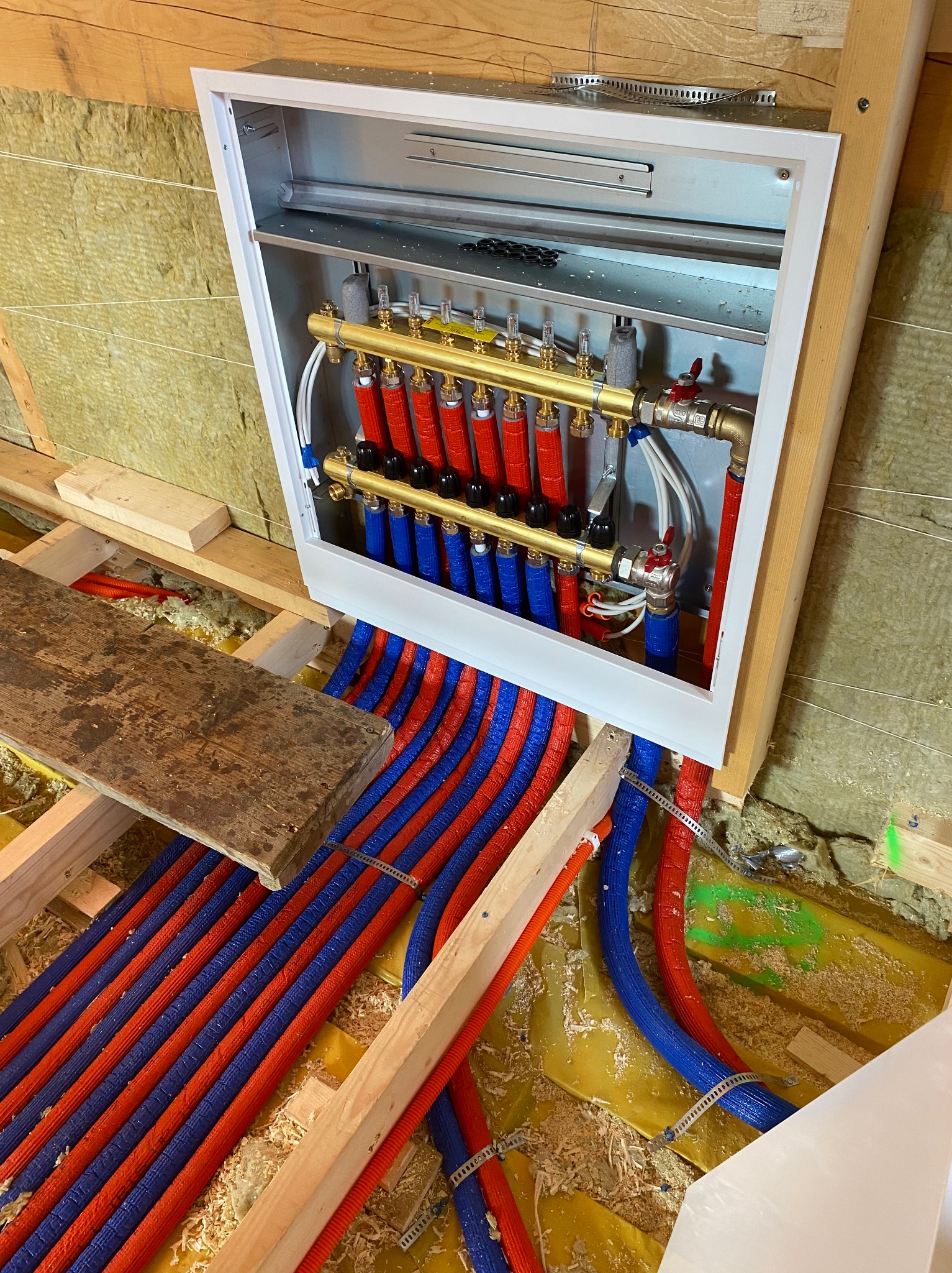 Rozdzielacz ogrzewania podłogowego w białej obudowie, z widocznymi rurami w kolorach czerwonym i niebieskim, zamocowany w konstrukcji drewnianej z izolacją.