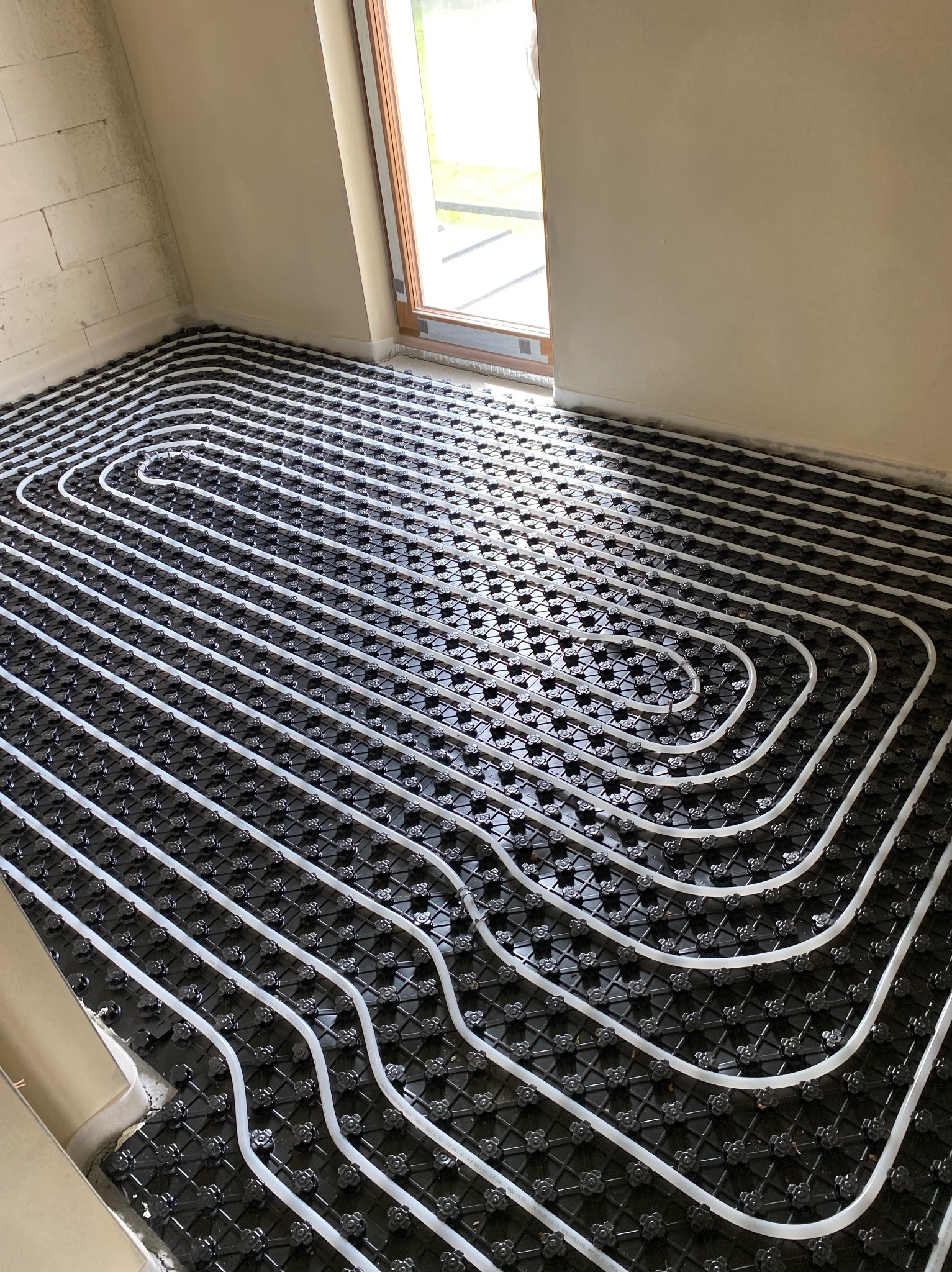 Białe rury ogrzewania podłogowego ułożone na czarnej macie w jasnym pomieszczeniu z oknem. Widok z góry, ukośny kadr.