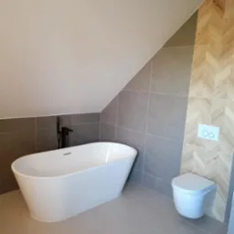 Nowoczesna łazienka na poddaszu z wolnostojącą białą wanną, szarą glazurą i drewnopodobną okładziną w jodełkę na ścianie, oraz podwieszaną toaletą.