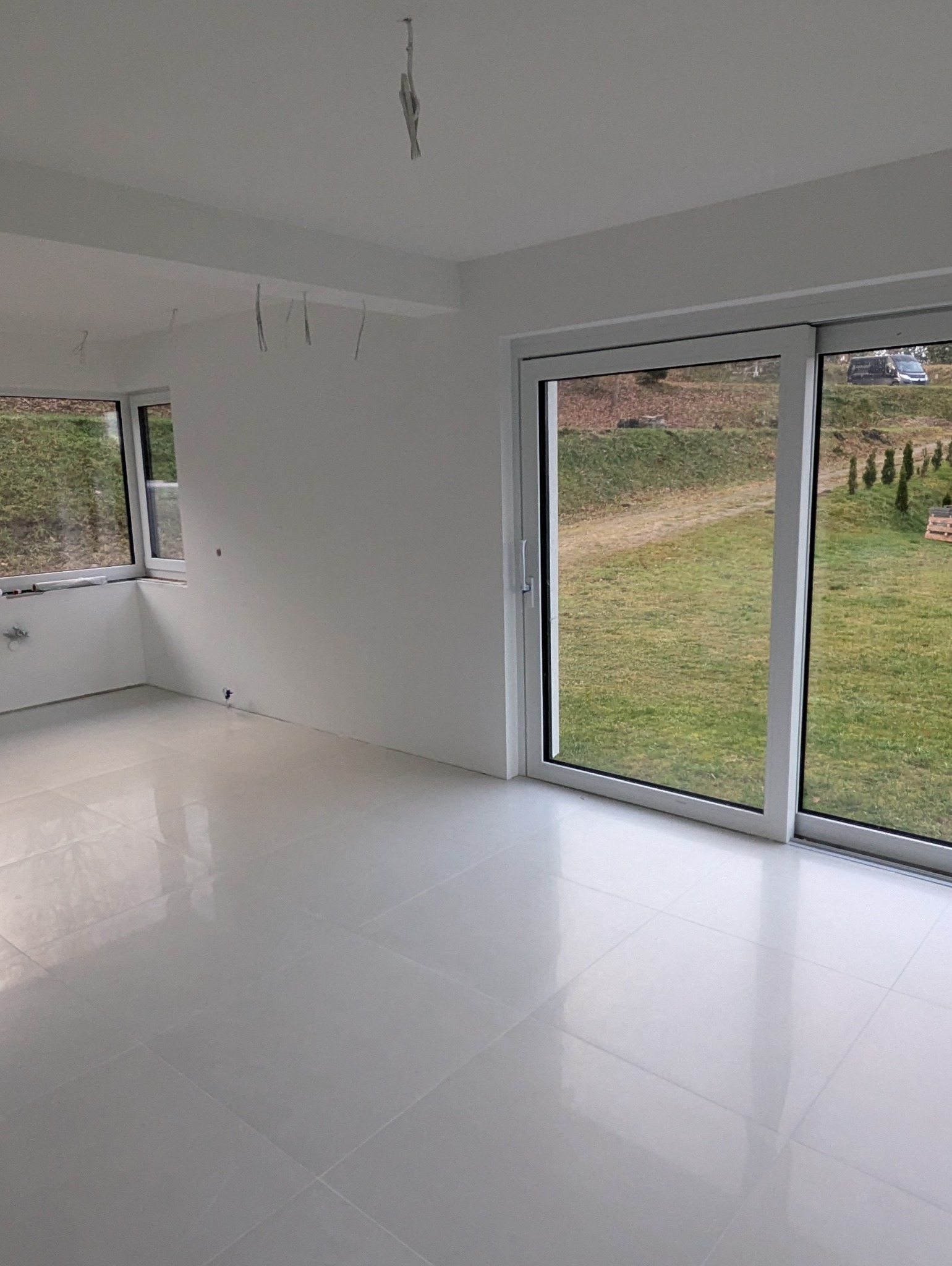 Białe, błyszczące płytki podłogowe w pustym, jasnym pokoju z dużym oknem przesuwnym i widocznymi kablami zwisającymi z sufitu.