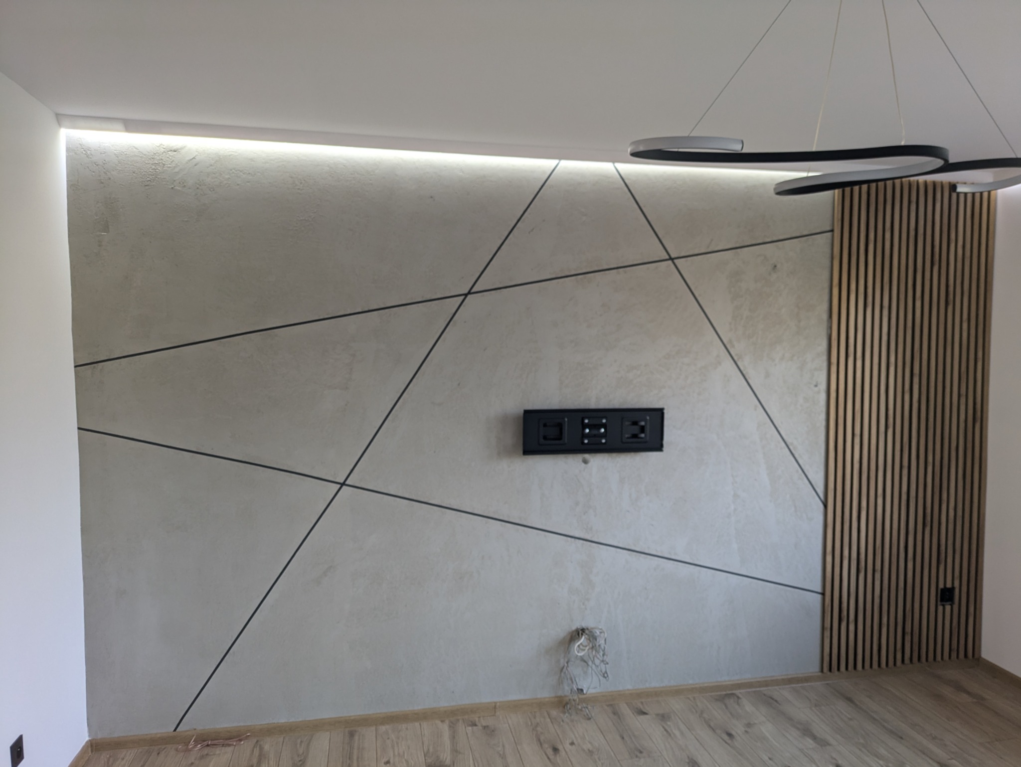 Ściana w salonie z dekoracyjną strukturą, podkreślona liniami tworzącymi geometryczny wzór, oświetlenie LED w górnej części, drewniane lamele po prawej stronie oraz uchwyt do telewizora; widoczne...