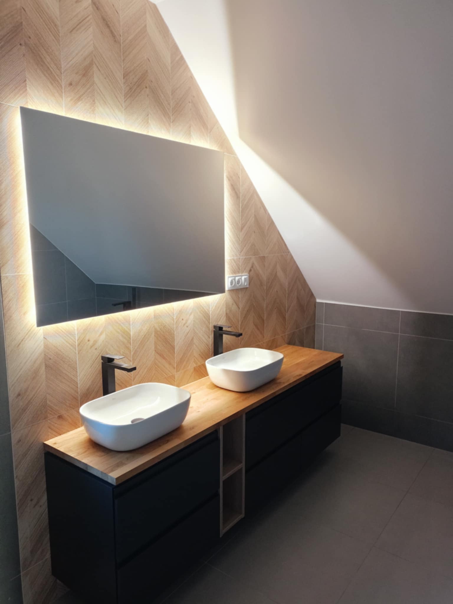 Nowoczesna łazienka na poddaszu z dwoma owalnymi umywalkami nablatowymi, czarną armaturą i podświetlanym lustrem na ścianie wyłożonej płytkami w jodełkę, szare płytki podłogowe i na części ściany.