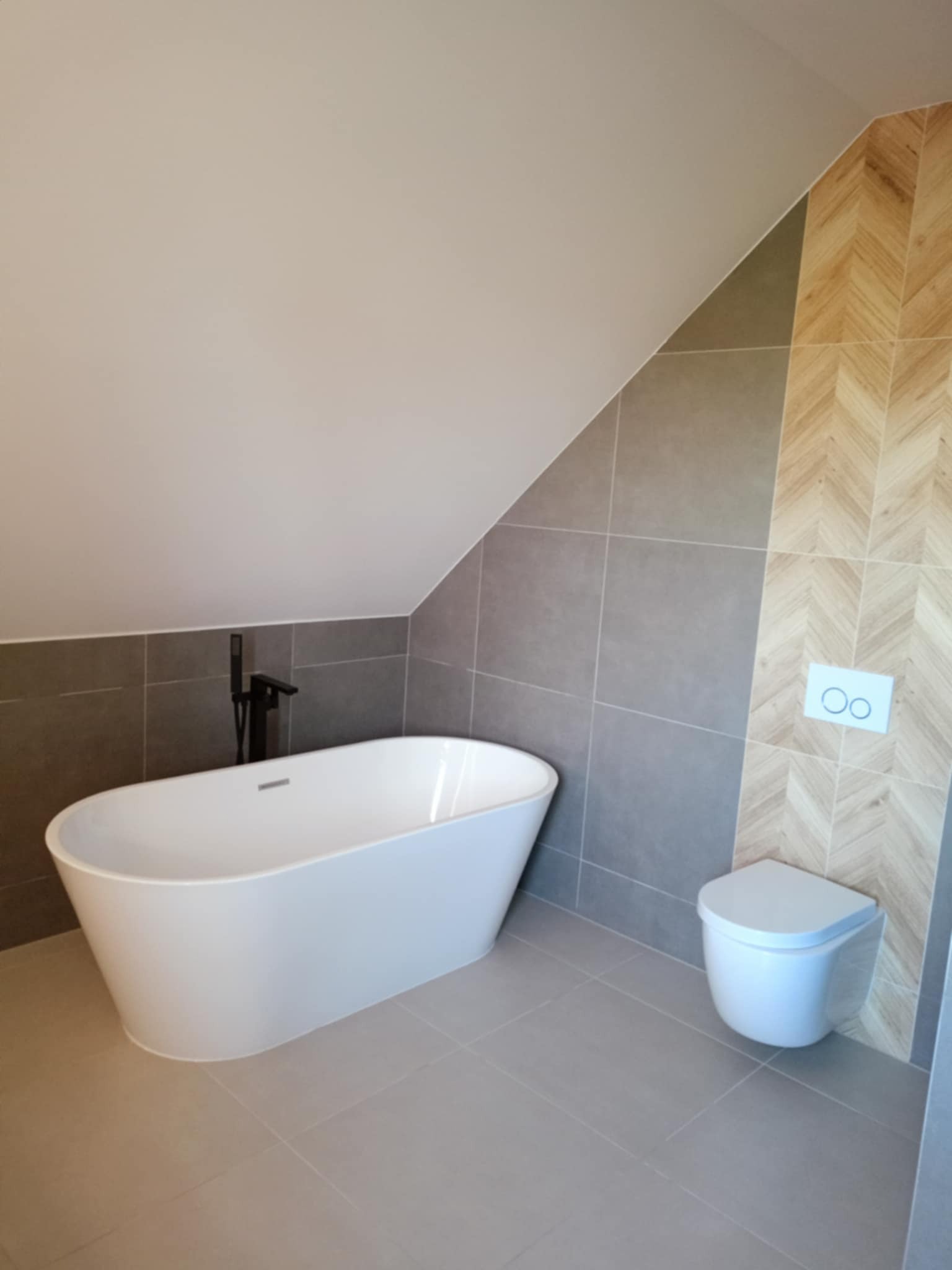 Nowoczesna łazienka na poddaszu z wolnostojącą białą wanną, szarą glazurą i drewnopodobną okładziną w jodełkę na ścianie, oraz podwieszaną toaletą.