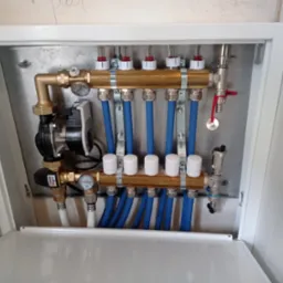 Rozdzielacz ogrzewania podłogowego w metalowej szafce, widoczne manometry, zawory, niebieskie rury i pompa obiegowa ESBE.