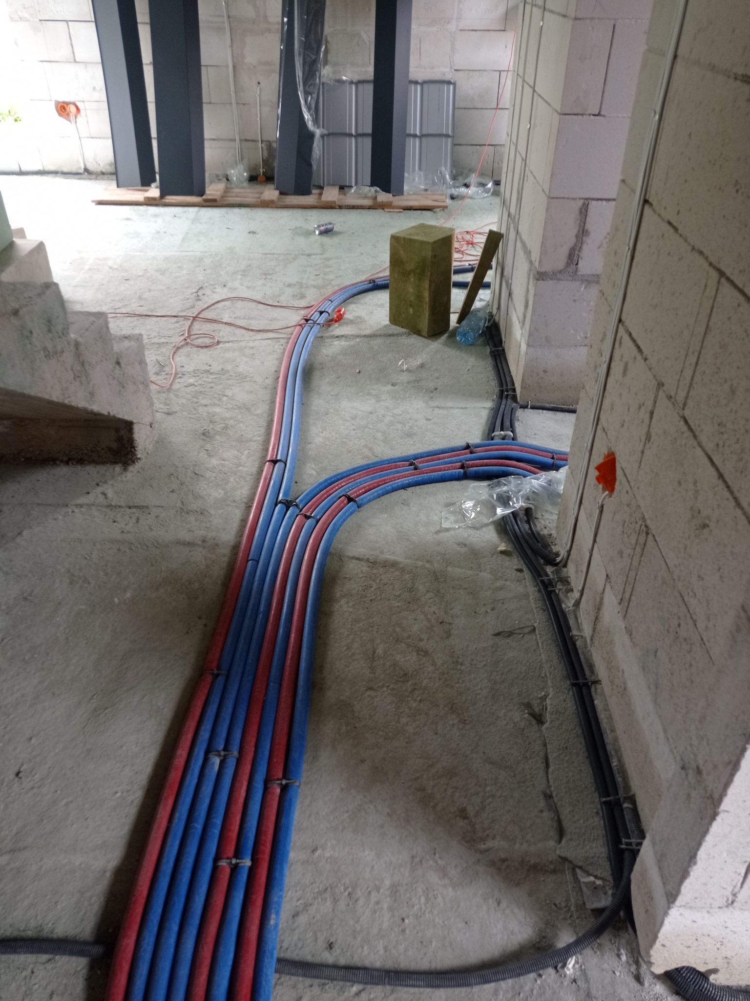 Instalacja rur grzewczych i wodnych w surowym wnętrzu budynku, rury prowadzone po podłodze i mocowane do ściany z betonu komórkowego, widoczna kostka izolacyjna i puszka elektryczna.