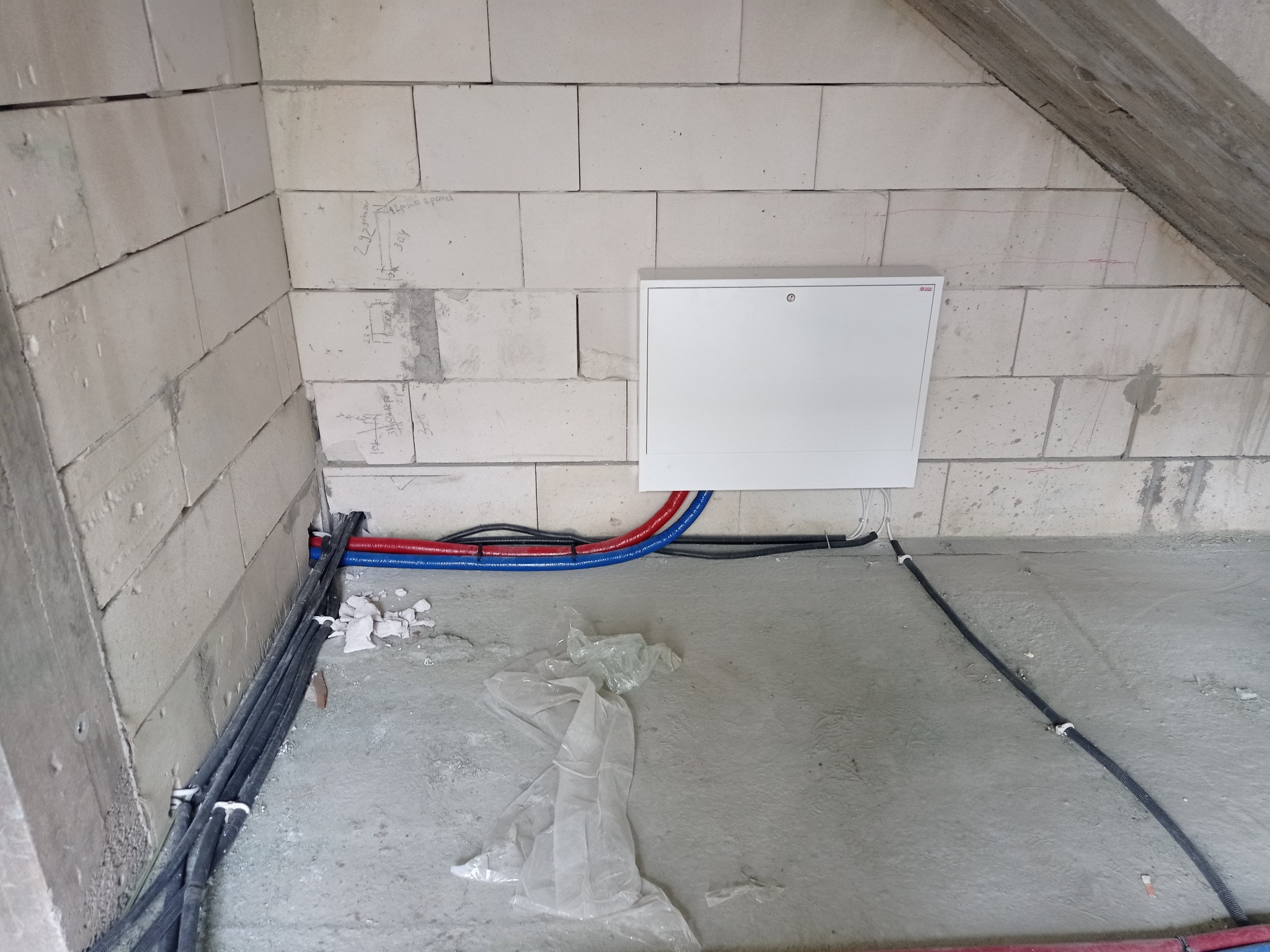 Surowy stan: Instalacja rur grzewczych (czerwone i niebieskie) oraz przewodów elektrycznych w ścianie z bloczków betonowych, z białą skrzynką rozdzielczą i widocznymi notatkami na ścianie.