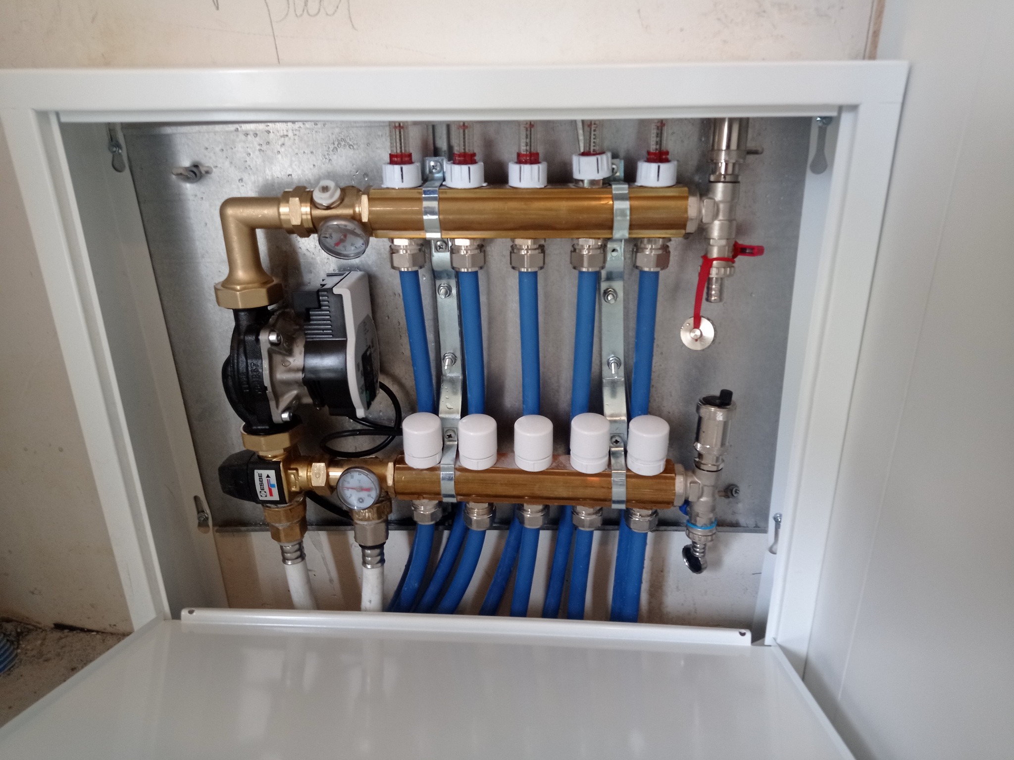 Rozdzielacz ogrzewania podłogowego w metalowej szafce, widoczne manometry, zawory, niebieskie rury i pompa obiegowa ESBE.