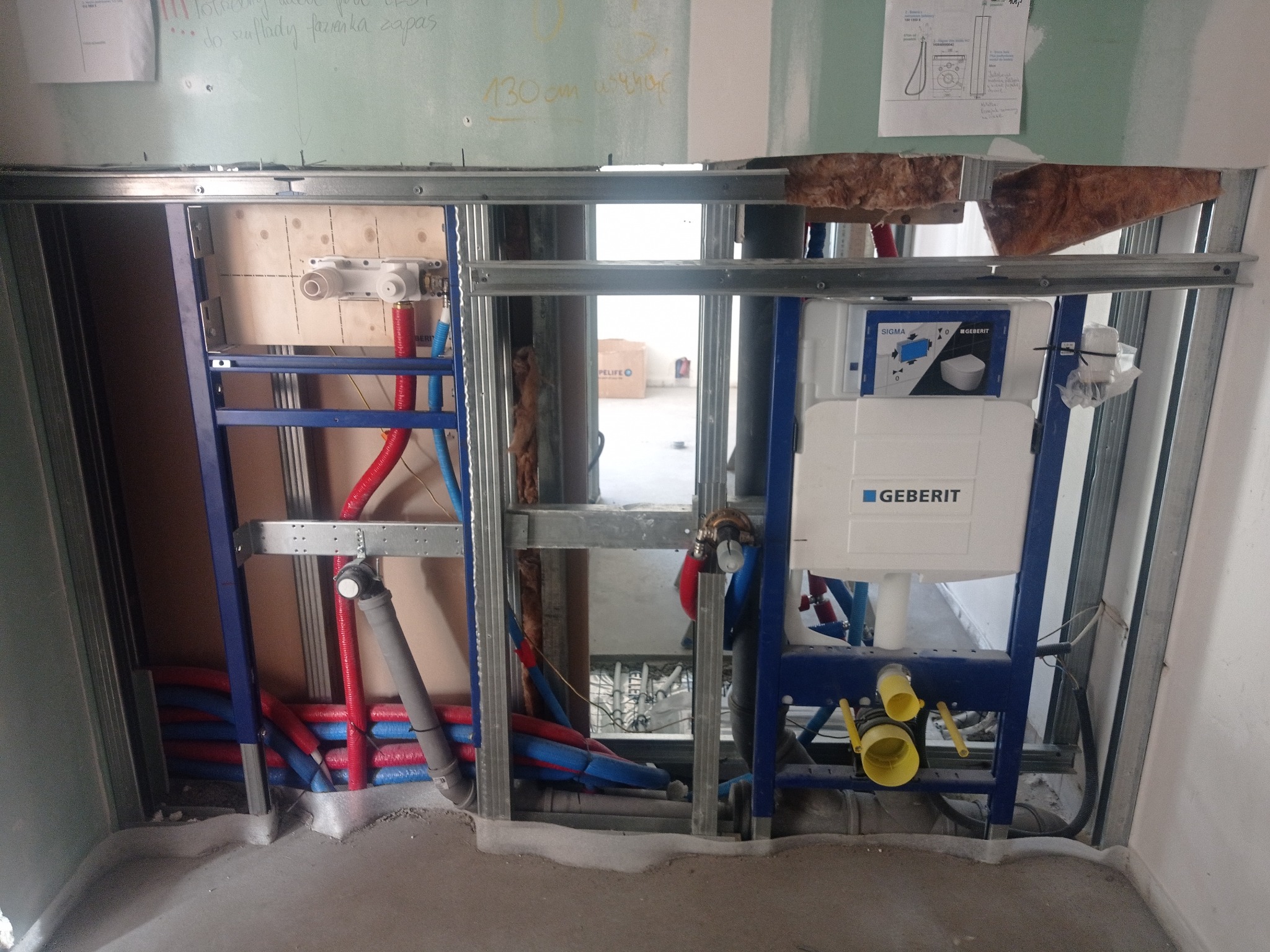 Stelaż podtynkowy Geberit z widocznymi rurami instalacji wodnej (czerwone i niebieskie) oraz kanalizacyjnej (szare) w trakcie montażu, częściowo zabudowany płytą gipsowo-kartonową, z widocznymi...