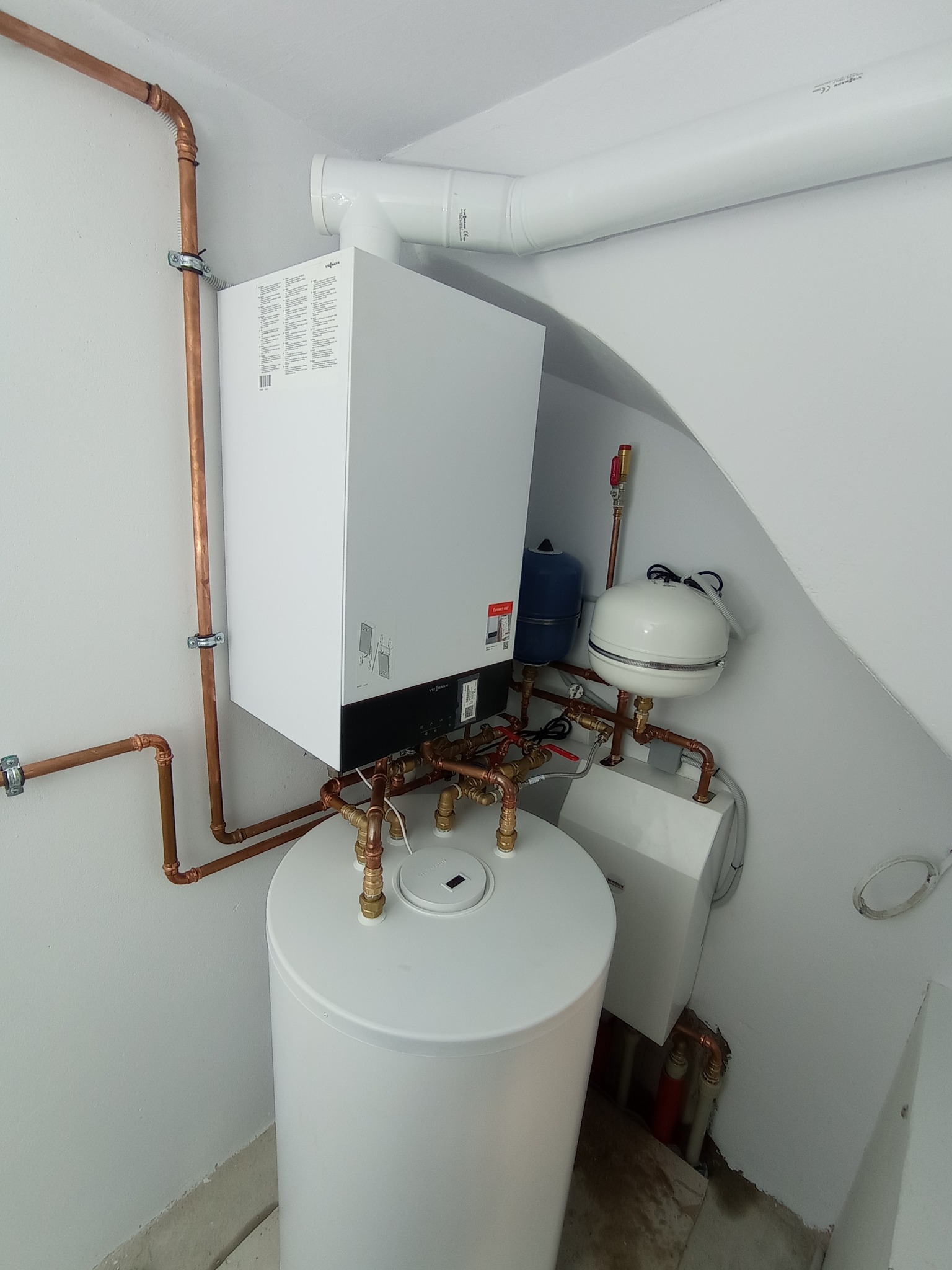 Nowoczesny, biały piec CO marki Viessmann z zasobnikiem wody, widoczne miedziane rury instalacji grzewczej i białe elementy wentylacji w jasnym pomieszczeniu kotłowni.