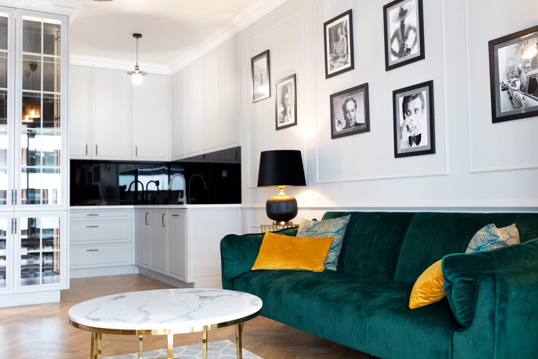 Salon z aneksem kuchennym w stylu glamour: szmaragdowa sofa, marmurowy stolik kawowy na złotych nogach, ściana z czarno-białymi portretami, biała zabudowa kuchenna z czarnym lacobelem.