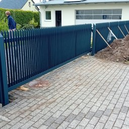 Firma Usługowo Budowlana Leszek Butylinski - Nowoczesne, ciemnoniebieskie ogrodzenie metalowe z pionowymi prętami o ostrych zakończeniach, widoczne podczas montażu, z pryzmą ziemi obok i chodnikiem z kostki brukowej.
