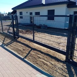 Firma Usługowo Budowlana Leszek Butylinski - Nowoczesne ogrodzenie panelowe w kolorze grafitowym, z przesuwną bramą, na tle jasnego domu z ciemnym dachem i kostki brukowej. Słoneczny dzień, cień ogrodzenia na chodniku.