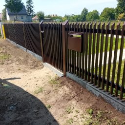 Metvis Leszek Butyliński - Nowoczesne, brązowe ogrodzenie metalowe z furtką i skrzynką na listy, zamontowane na betonowej podmurówce. Widok na posesję z zewnątrz. Słoneczny dzień.