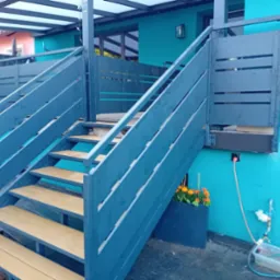 Nowoczesne, pomalowane na niebiesko schody zewnętrzne z drewnianymi stopniami, prowadzące na taras domu o turkusowej elewacji. Widoczna zieleń i instalacja nawadniająca.