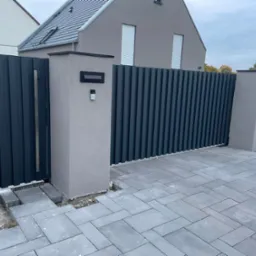 Nowoczesna, grafitowa brama wjazdowa z pionowych paneli, osadzona w szarych, murowanych słupkach. Dojście wyłożone szarą kostką brukową. Tło stanowi szary dom z ciemnym dachem.