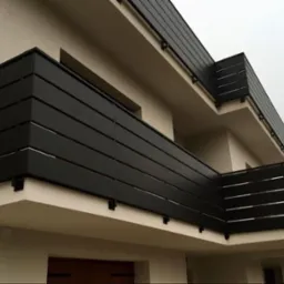 Nowoczesna balustrada balkonowa z poziomych, czarnych paneli, zamontowana na jasnym budynku. Minimalistyczny design, widok z dołu, pochmurne niebo w tle.