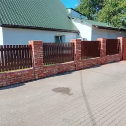 Brązowe, drewniane ogrodzenie z ceglanymi słupkami na tle białego domu z zielonym dachem, widziane z poziomu chodnika z kostki brukowej.