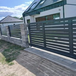 Metvis Leszek Butyliński - Nowoczesne, ciemnoszare ogrodzenie z poziomymi lamelami i przesuwną bramą, wsparte na słupkach z bloczków betonowych. W tle biały dom z numerem 70b.