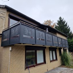 Metvis Leszek Butyliński - Nowoczesna, ciemnoszara balustrada balkonowa z metalu, ozdobiona laserowo wyciętym wzorem stylizowanych traw i cegieł, z nierdzewną poręczą, zamontowana na jasnym ceglanym domu.
