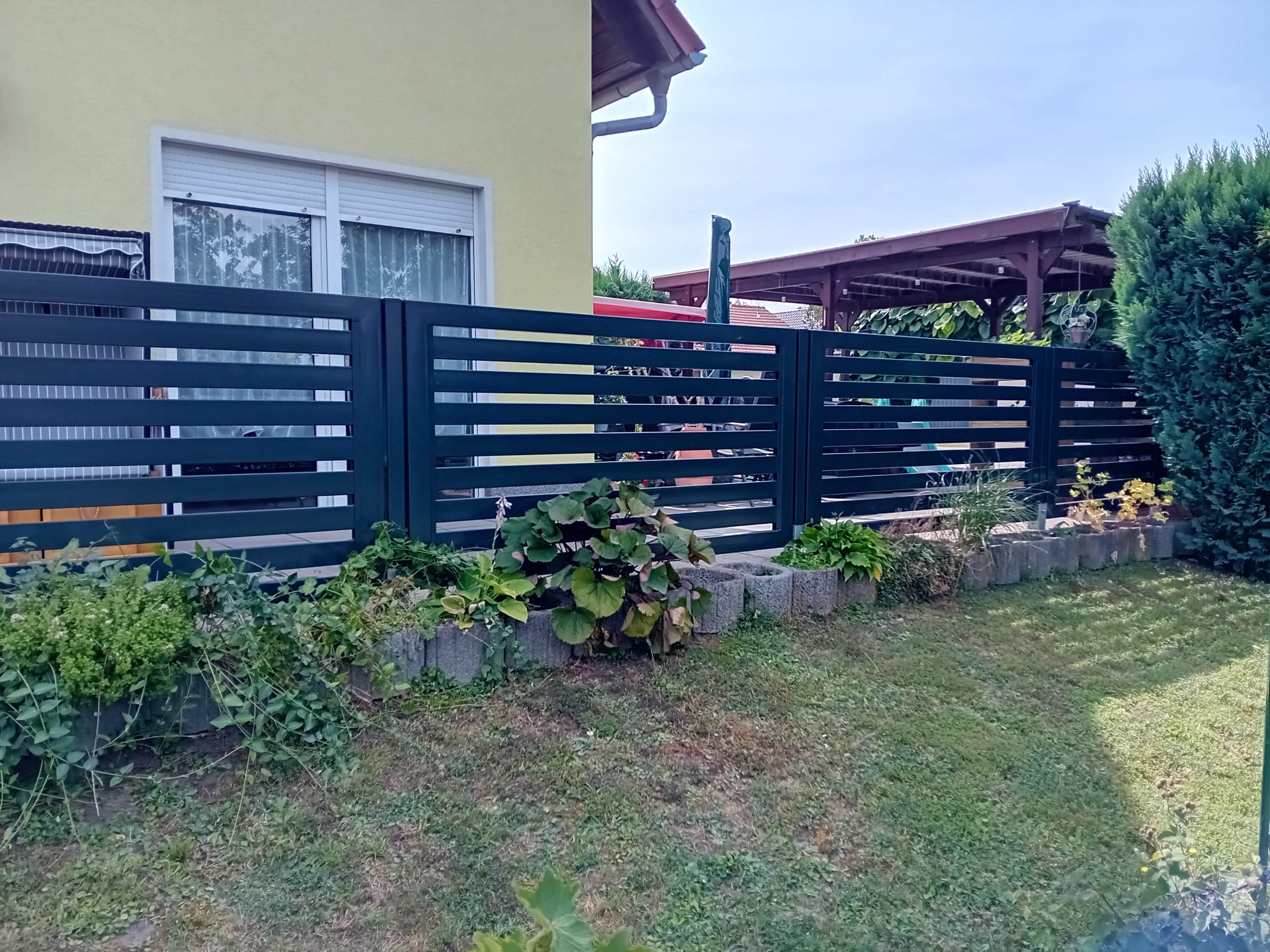 Nowoczesne, poziome ogrodzenie panelowe w kolorze grafitowym, z widoczną konstrukcją domu w tle i roślinnością wzdłuż ogrodzenia.