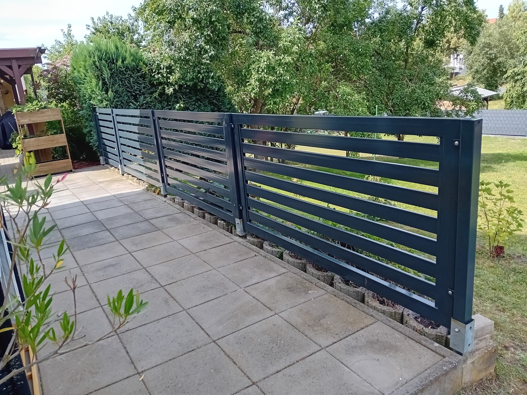 Nowoczesne, poziome przęsła ogrodzenia z metalu w kolorze grafitowym, zamontowane na niskiej podmurówce z betonowych elementów, widok z perspektywy tarasu wyłożonego szarą kostką brukową.