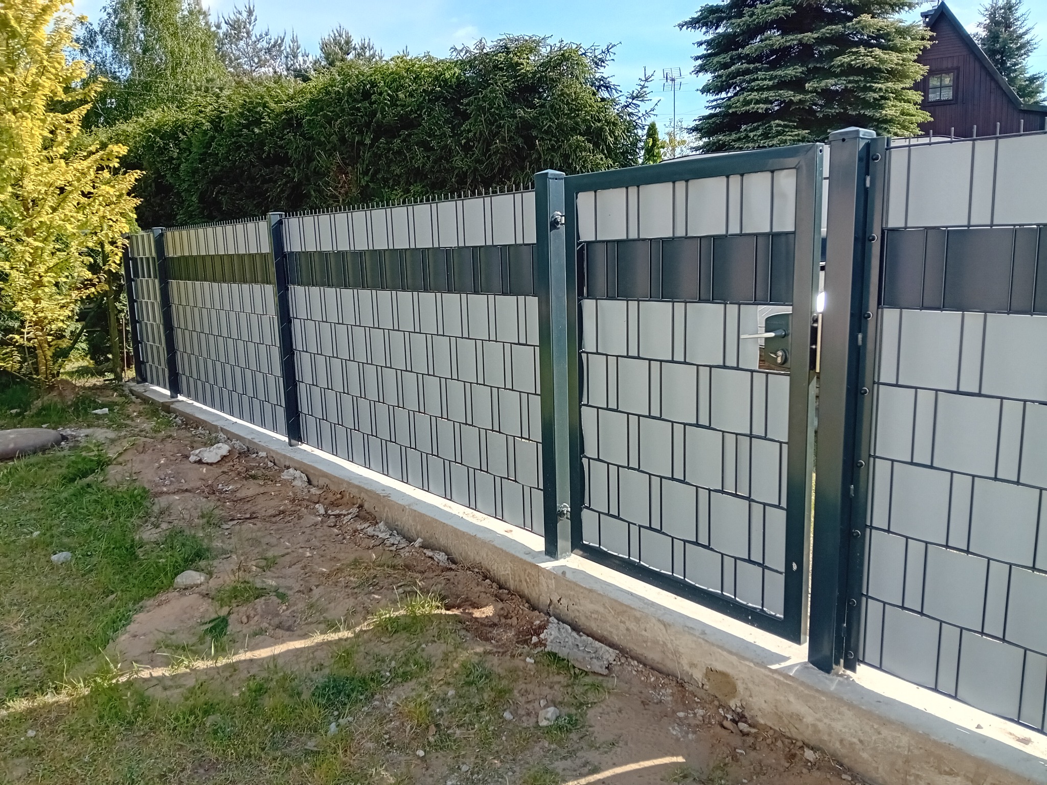 Nowoczesne ogrodzenie panelowe z bramką, wypełnione płytami w odcieniach szarości i grafitu, osadzone na betonowej podmurówce, na tle zieleni.