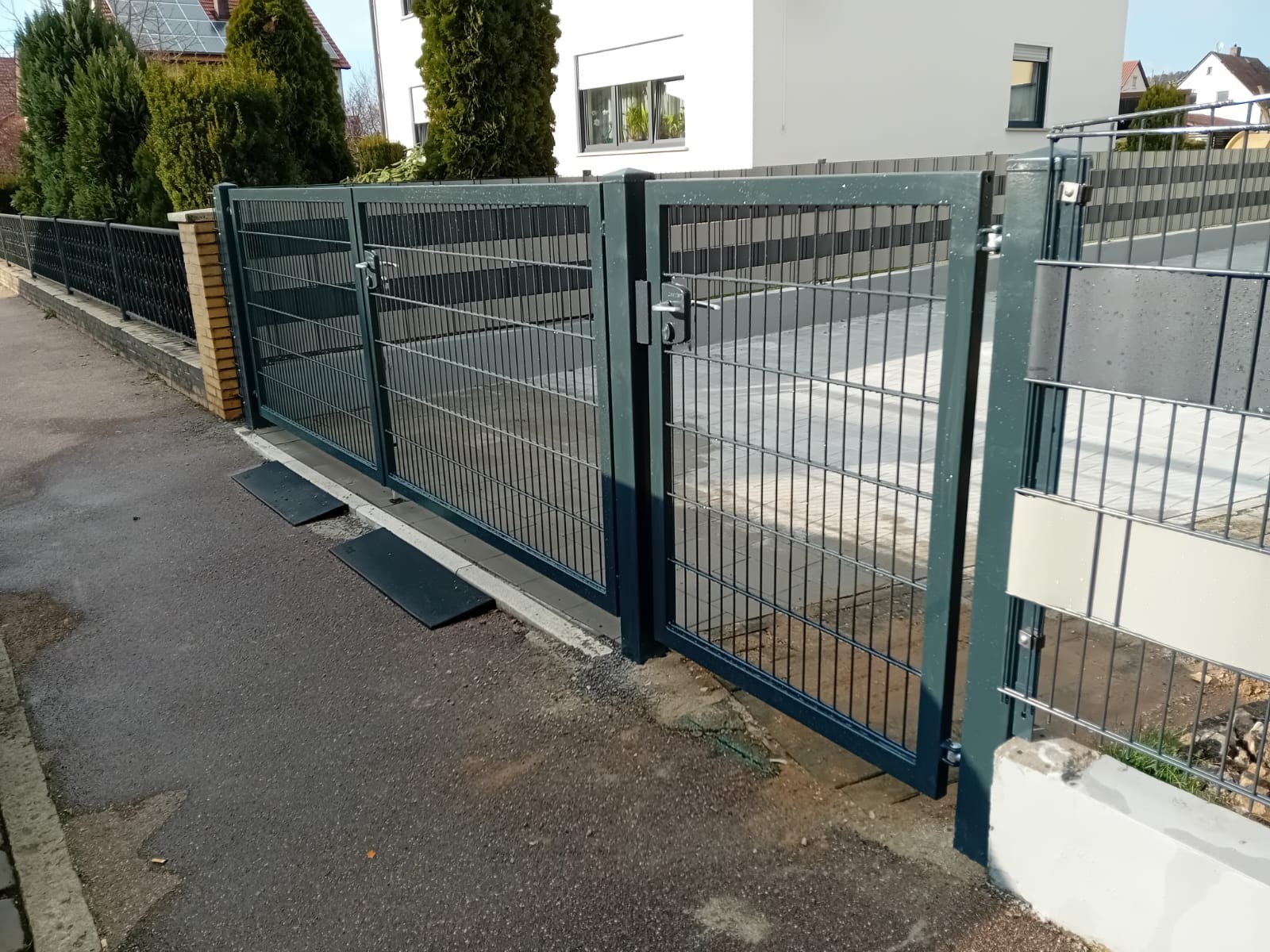 Otwarta, nowoczesna brama panelowa w kolorze grafitowym, z widocznymi detalami zamka i słupków. W tle dom z białymi ścianami i fragment ogrodzenia panelowego.