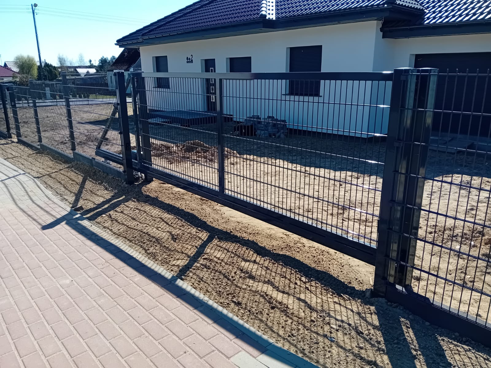 Nowoczesne ogrodzenie panelowe w kolorze grafitowym, z przesuwną bramą, na tle jasnego domu z ciemnym dachem i kostki brukowej. Słoneczny dzień, cień ogrodzenia na chodniku.