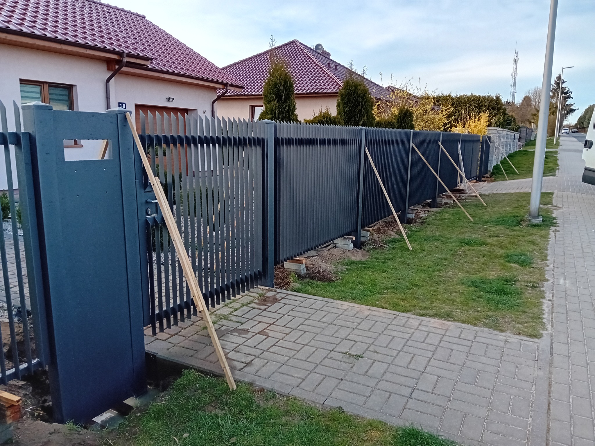 Ciemnoszary, nowoczesny płot panelowy w trakcie montażu, podparty drewnianymi belkami, widoczny na tle domów jednorodzinnych i chodnika z kostki.