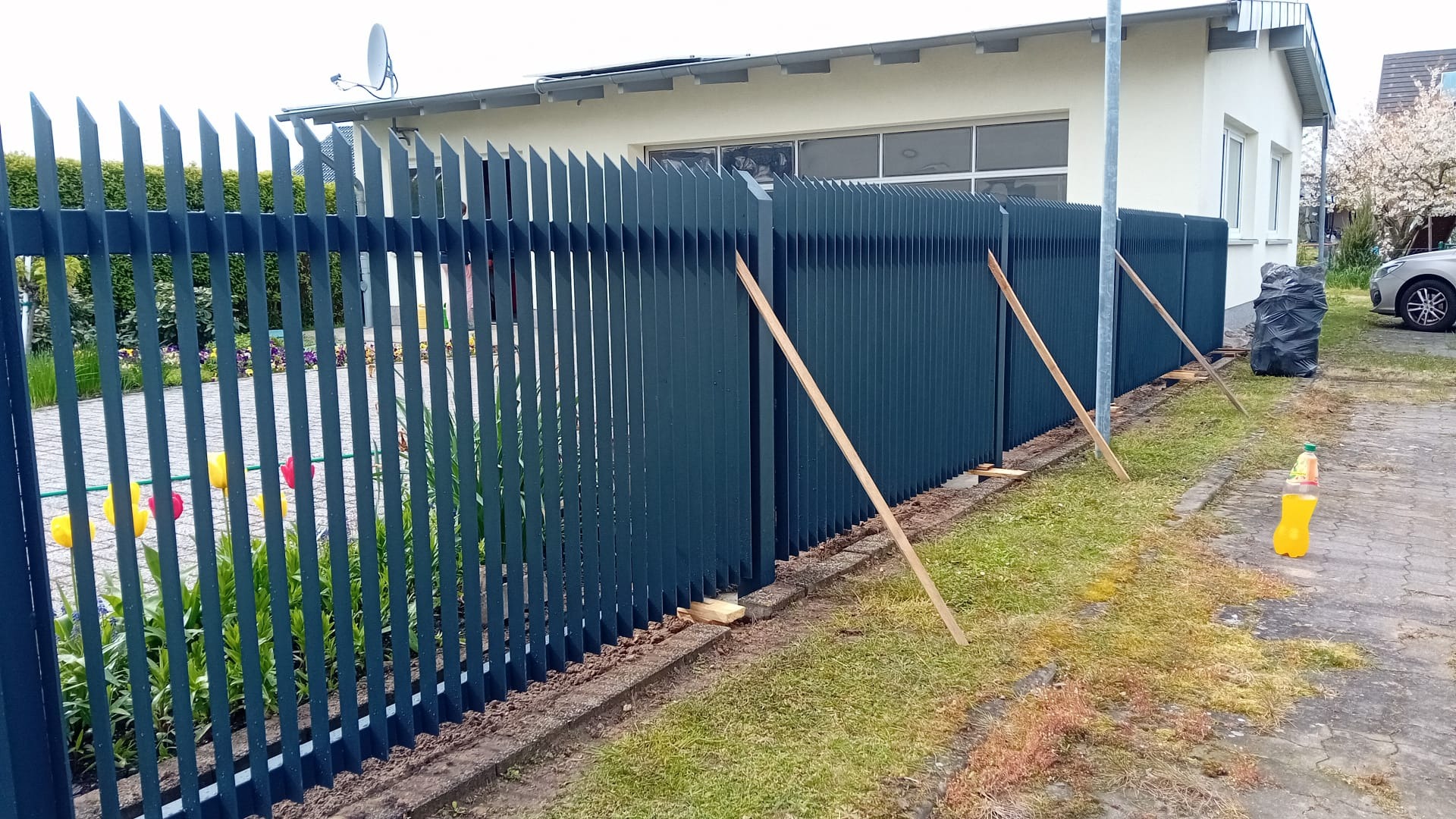 Nowoczesne, ciemnozielone ogrodzenie metalowe z ostrymi zakończeniami, częściowo zamontowane. Widoczne drewniane podpory i kwiaty w tle, dom z anteną satelitarną.