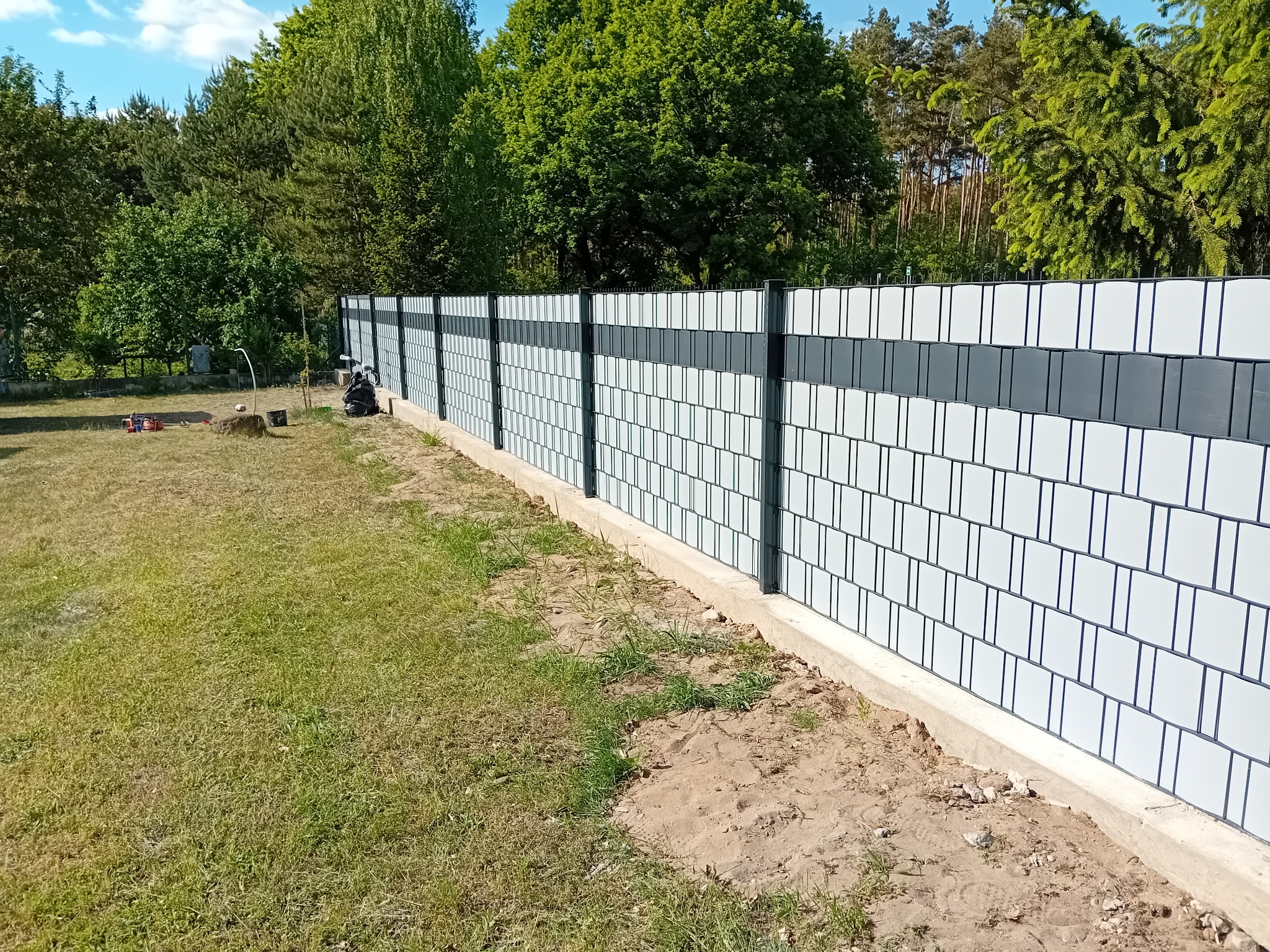 Nowoczesne ogrodzenie panelowe z białymi i szarymi wypełnieniami, widoczne narzędzia i częściowo sylwetki osób podczas montażu, trawnik i drzewa w tle.