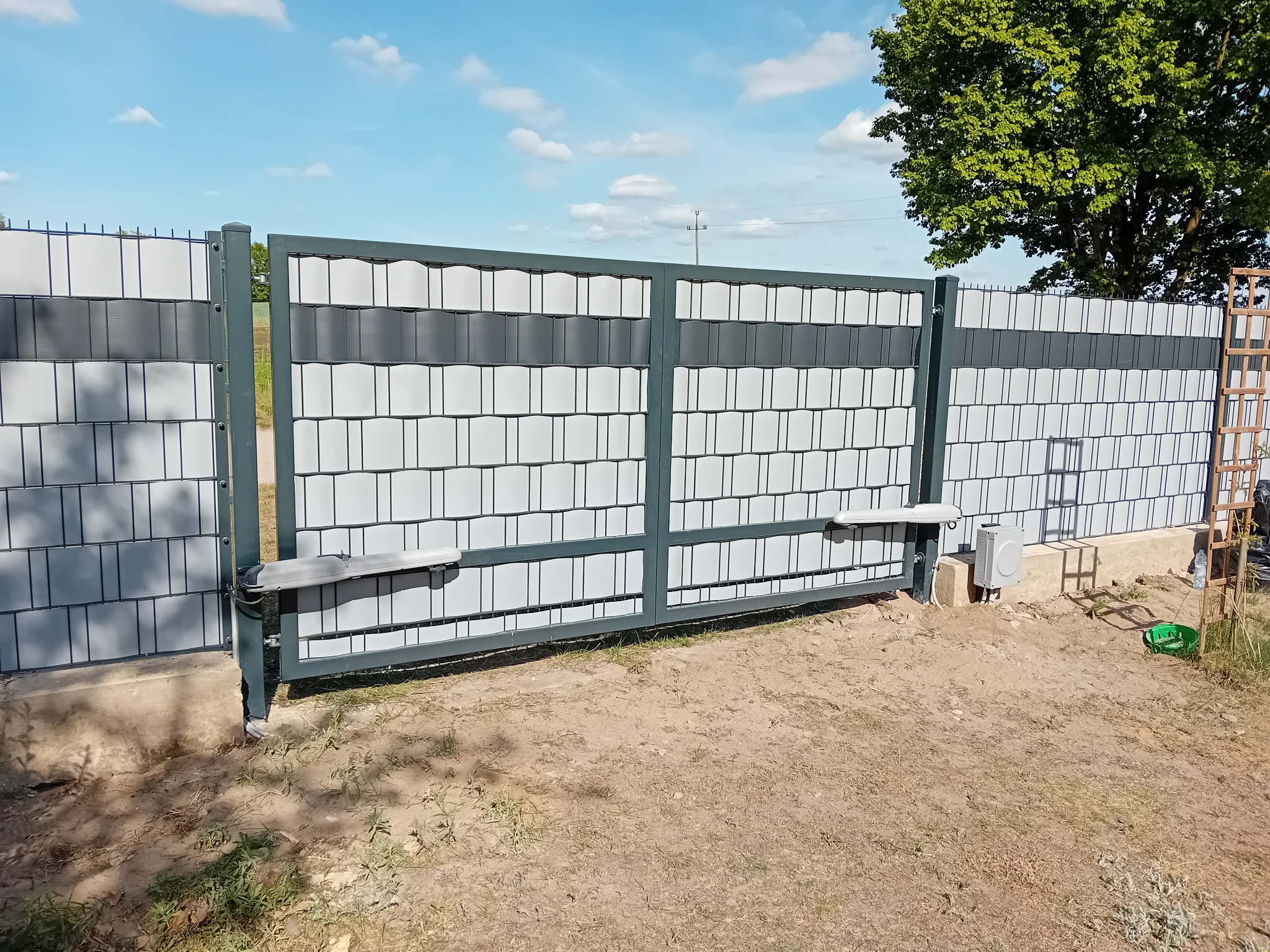 Nowoczesna, przesuwna brama panelowa z automatyką, wypełniona panelami w odcieniach szarości i bieli, na tle zieleni drzew i błękitnego nieba.