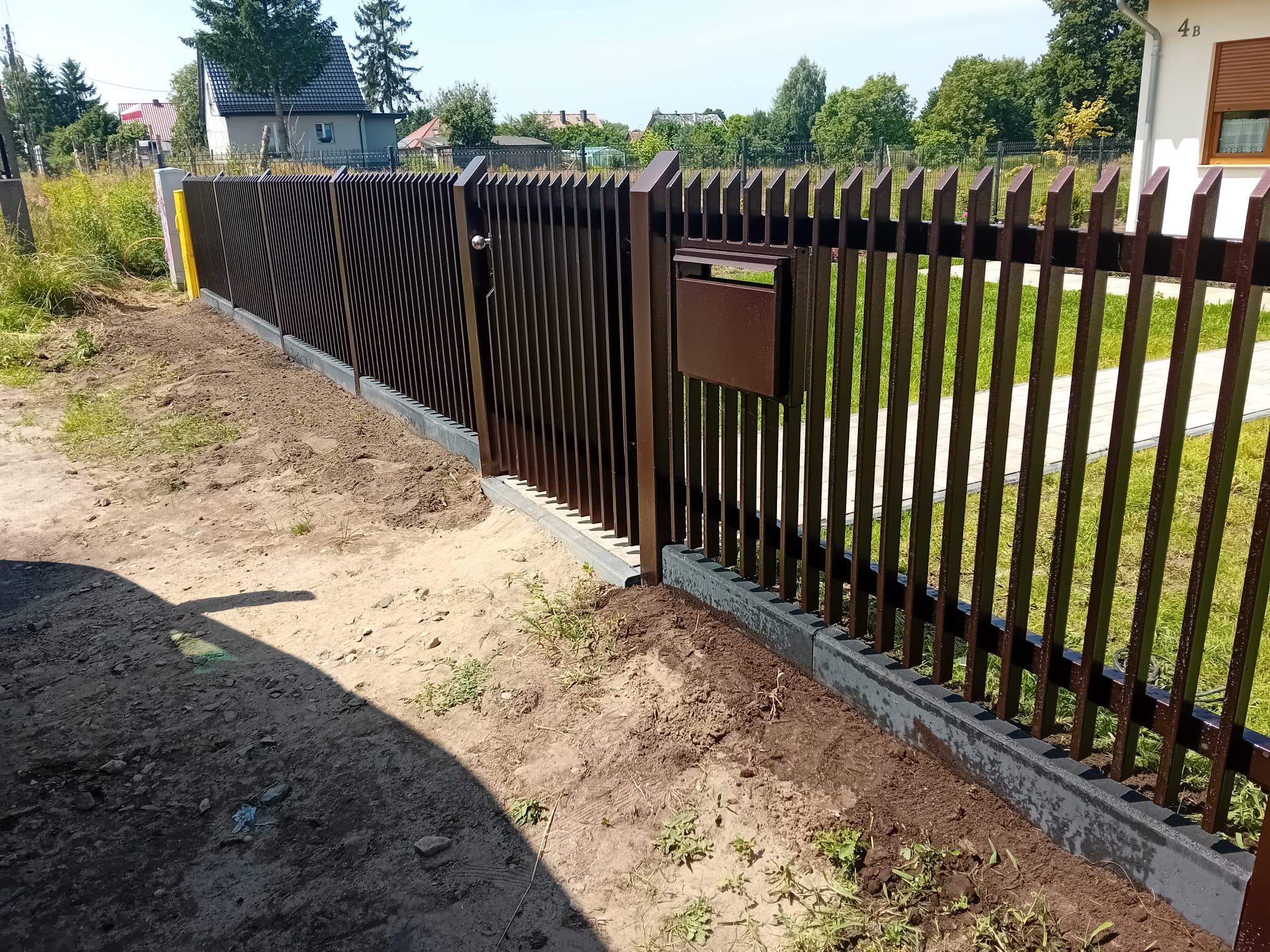 Nowoczesne, brązowe ogrodzenie metalowe z furtką i skrzynką na listy, zamontowane na betonowej podmurówce. Widok na posesję z zewnątrz. Słoneczny dzień.