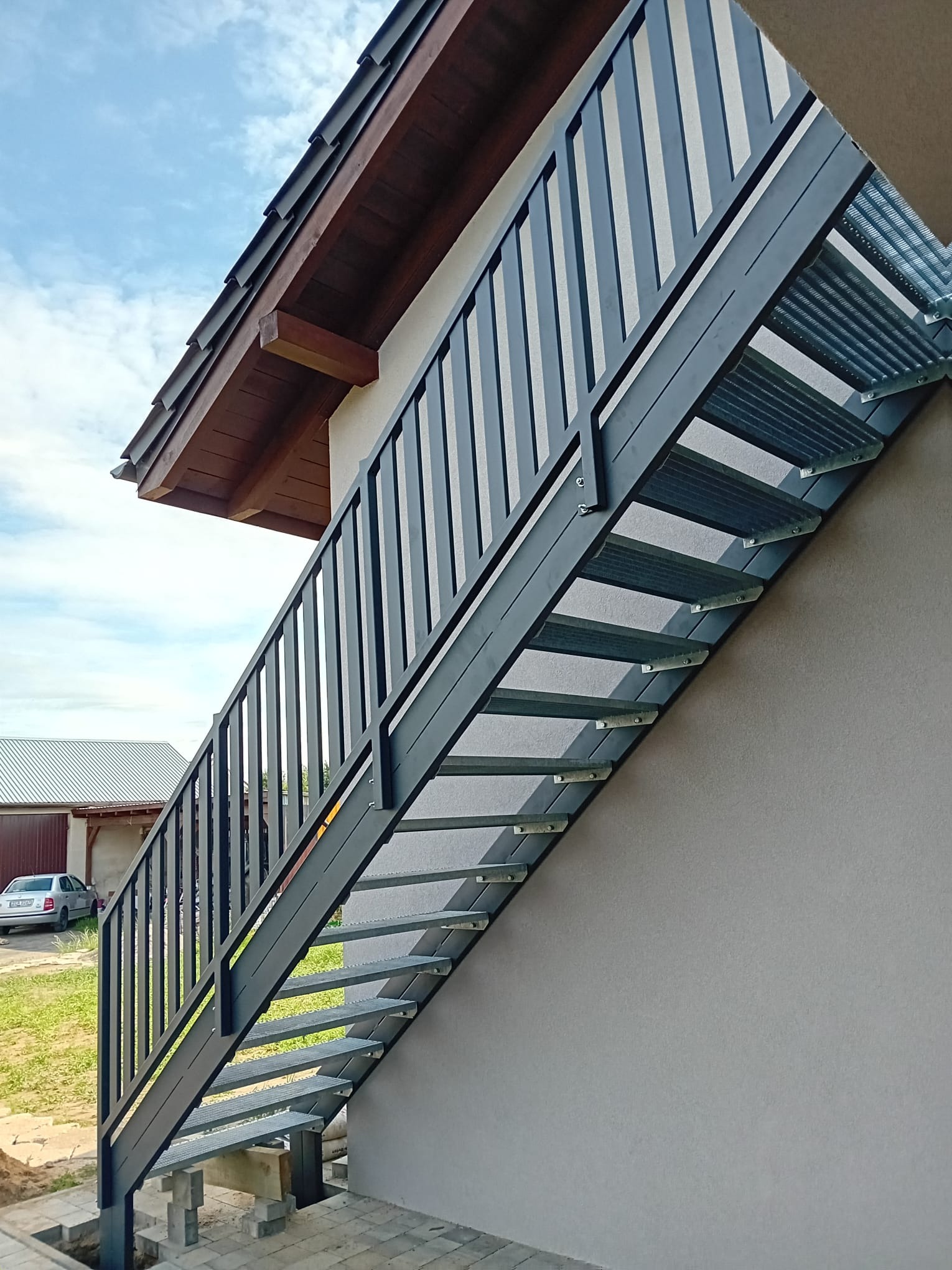 Zewnętrzne, metalowe schody z balustradą przymocowane do ściany budynku, widoczne z boku. Schody prowadzą w górę, w tle fragment dachu i zabudowań.