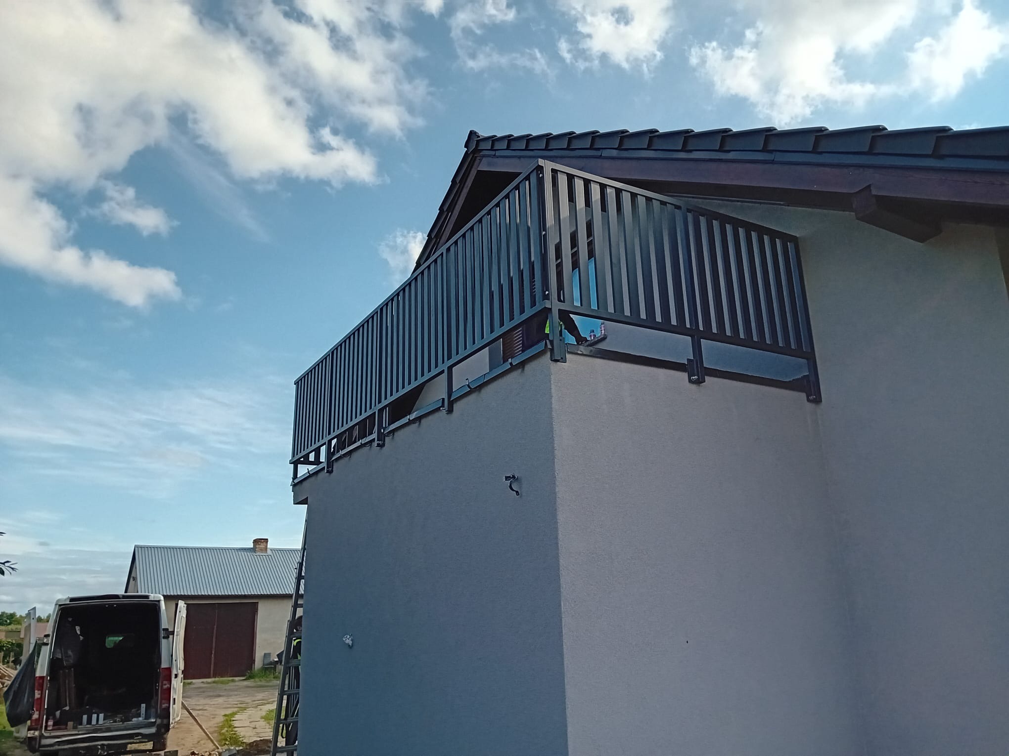 Czarna, metalowa balustrada balkonowa z pionowymi szczebelkami, zamontowana na rogu budynku. W tle błękitne niebo z chmurami i fragment dachu. Widoczny samochód dostawczy i drabina.