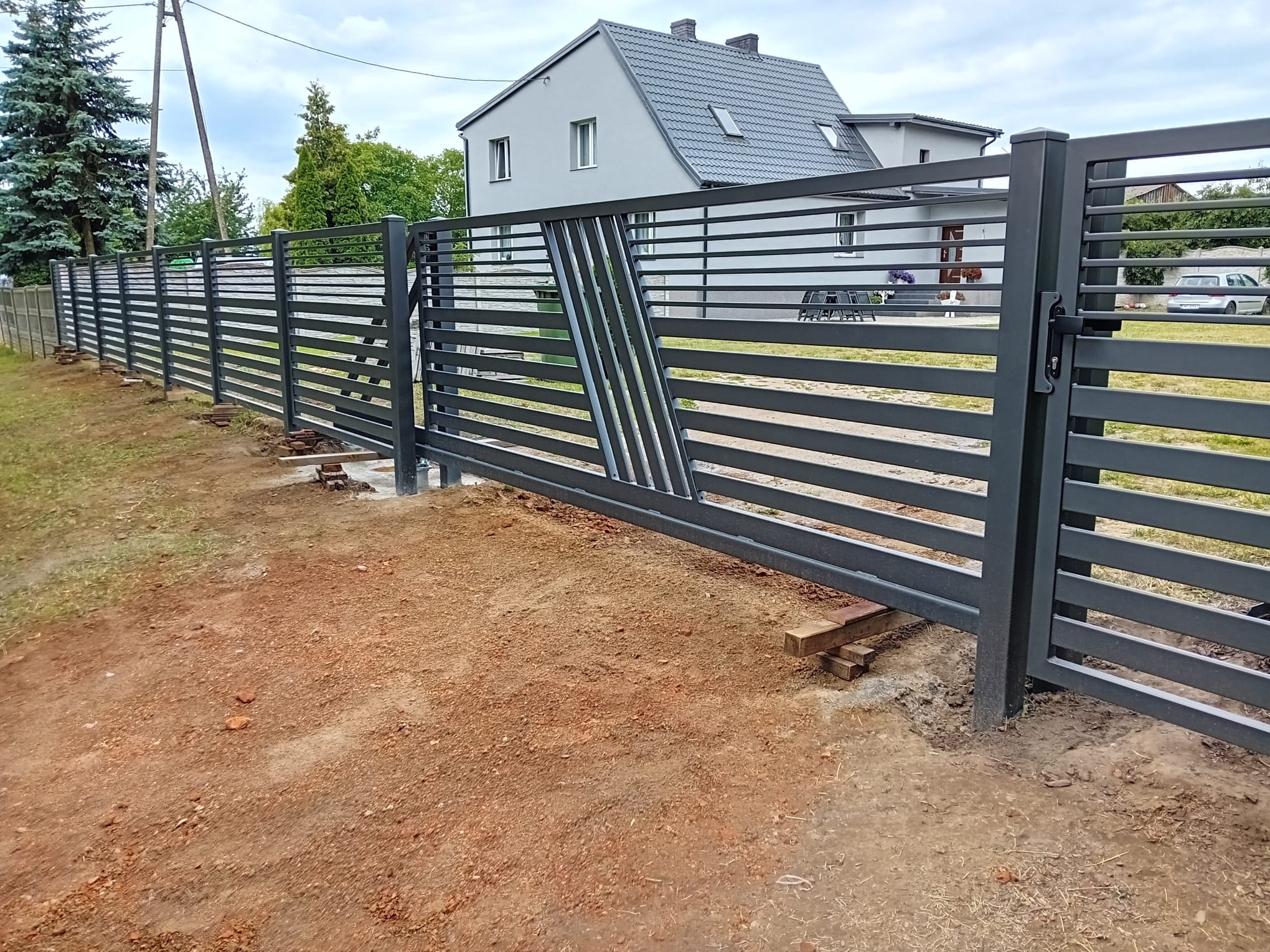 Nowoczesne, poziome przęsła metalowe w kolorze grafitowym tworzą ogrodzenie z bramą. Uwagę przykuwa ukośny wzór na bramie i surowy grunt wokół.