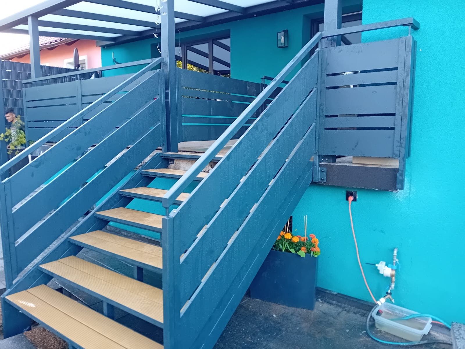 Nowoczesne, pomalowane na niebiesko schody zewnętrzne z drewnianymi stopniami, prowadzące na taras domu o turkusowej elewacji. Widoczna zieleń i instalacja nawadniająca.