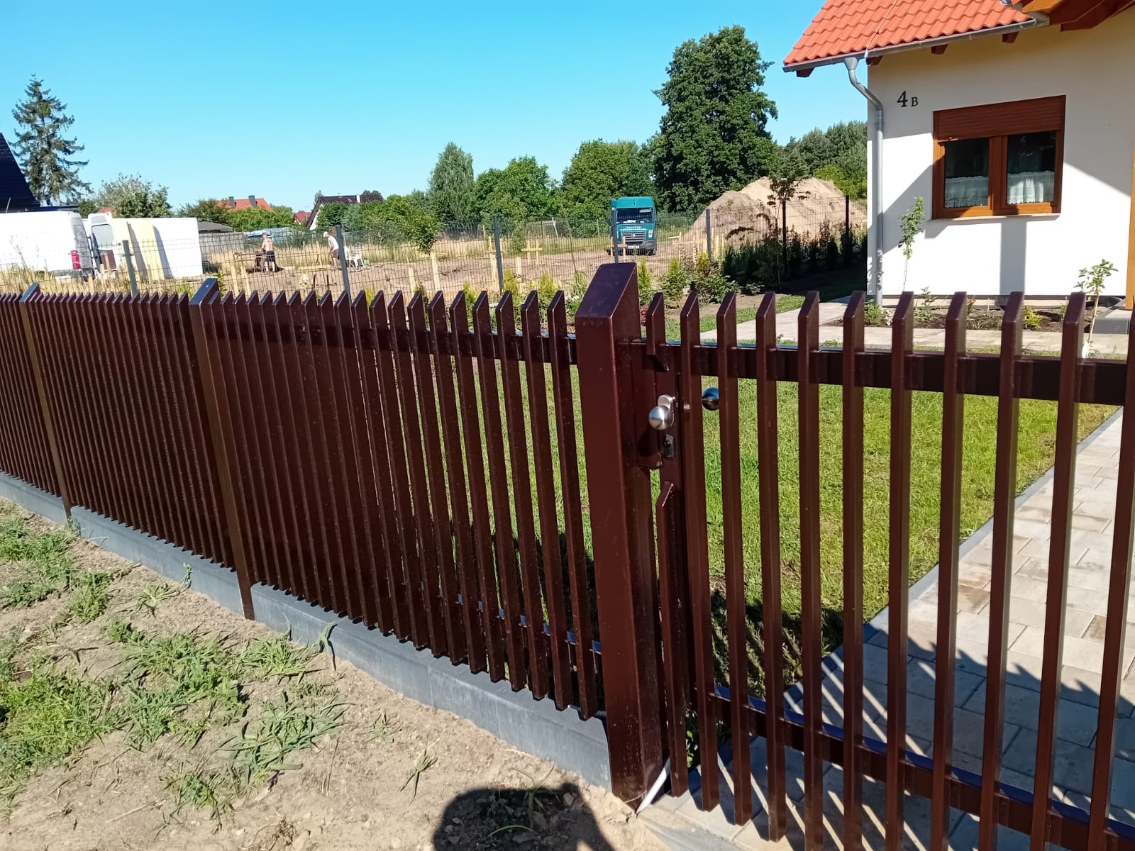 Brązowe, pionowe sztachety ogrodzenia metalowego z furtką i widocznym zamkiem na tle domu z ogrodem, w słoneczny dzień.