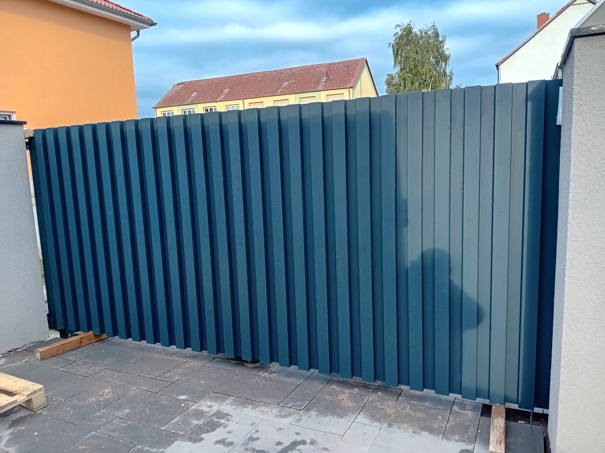 Nowoczesna, grafitowa brama przesuwna z pionowych paneli, zamontowana na posesji z kostki brukowej, z widocznym fragmentem budynku w tle.