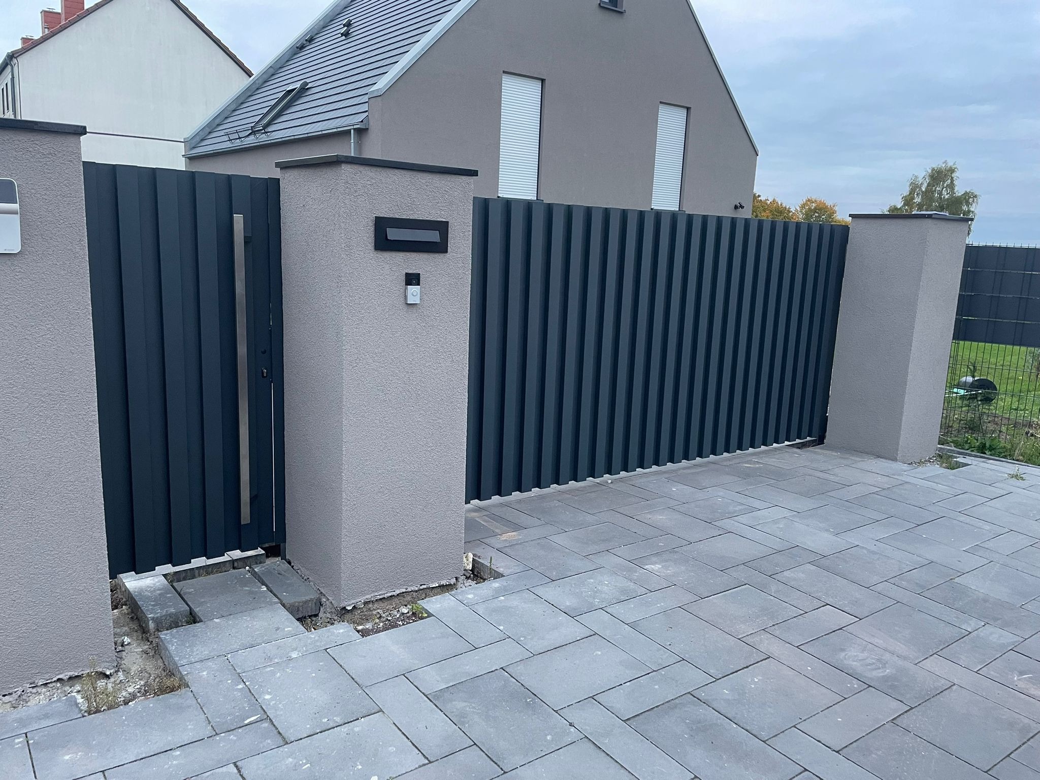 Nowoczesna, grafitowa brama wjazdowa z pionowych paneli, osadzona w szarych, murowanych słupkach. Dojście wyłożone szarą kostką brukową. Tło stanowi szary dom z ciemnym dachem.