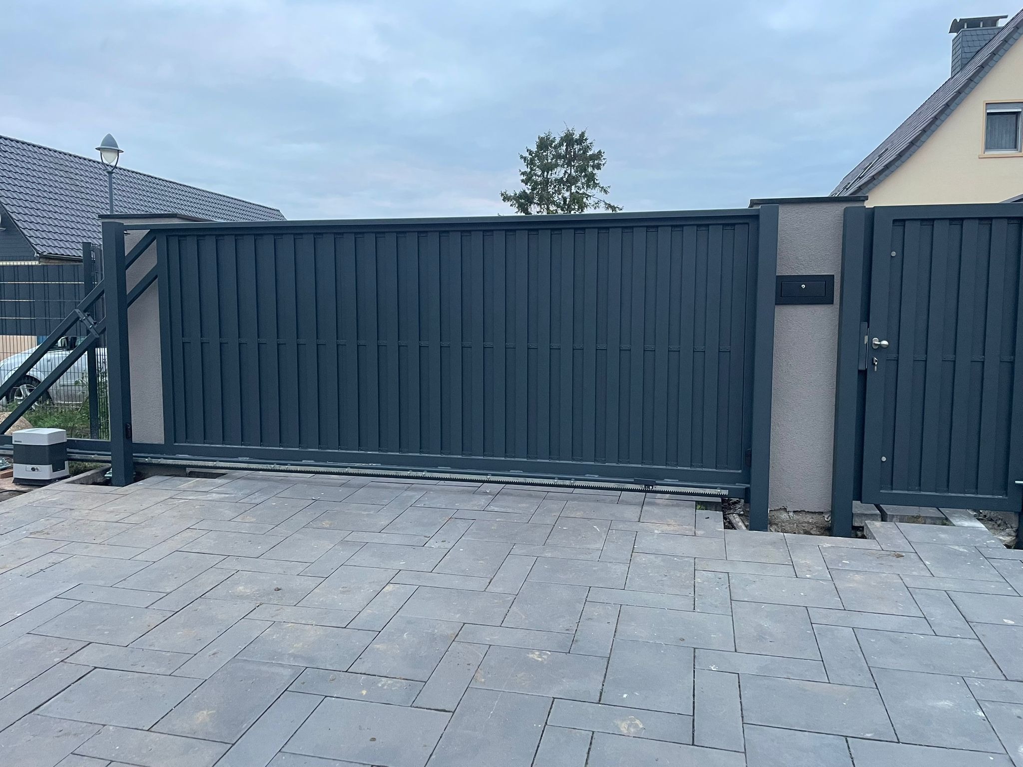 Nowoczesna, przesuwna brama wjazdowa z metalowymi panelami, zintegrowana ze słupkiem z szarą skrzynką pocztową, na tle kostki brukowej i domu.